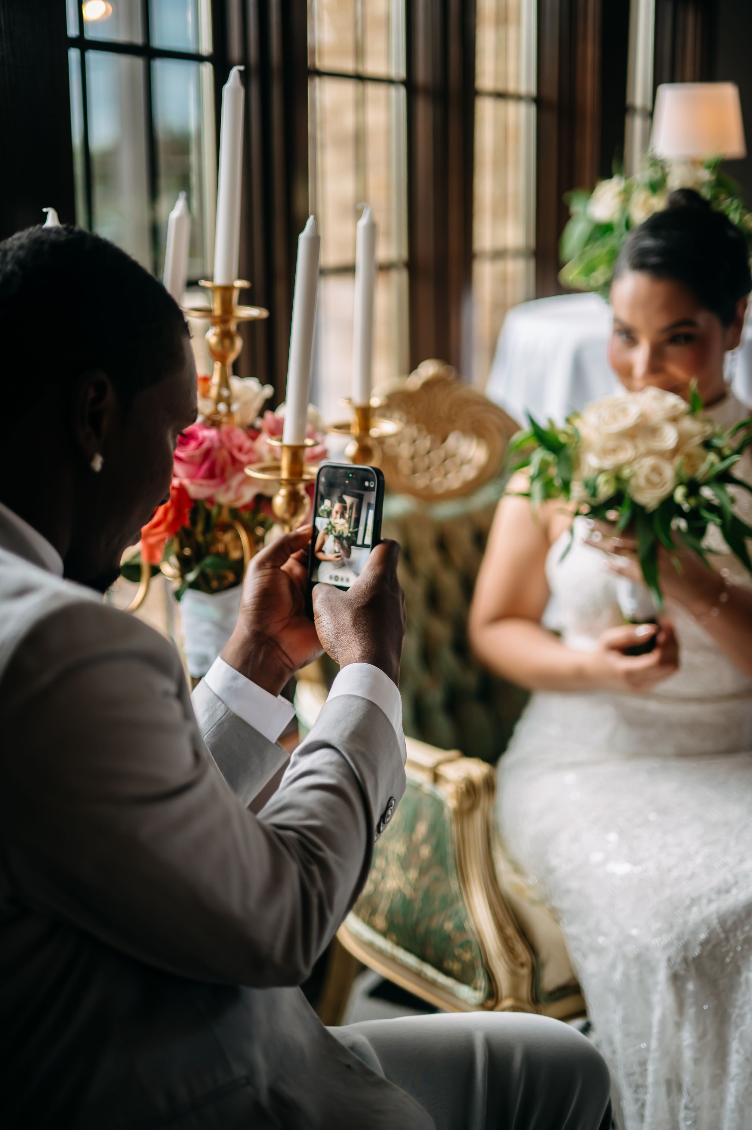 Groom in gray suit photographs bride holding bouquet on smartphone at elegant wedding reception with candles and flowers.