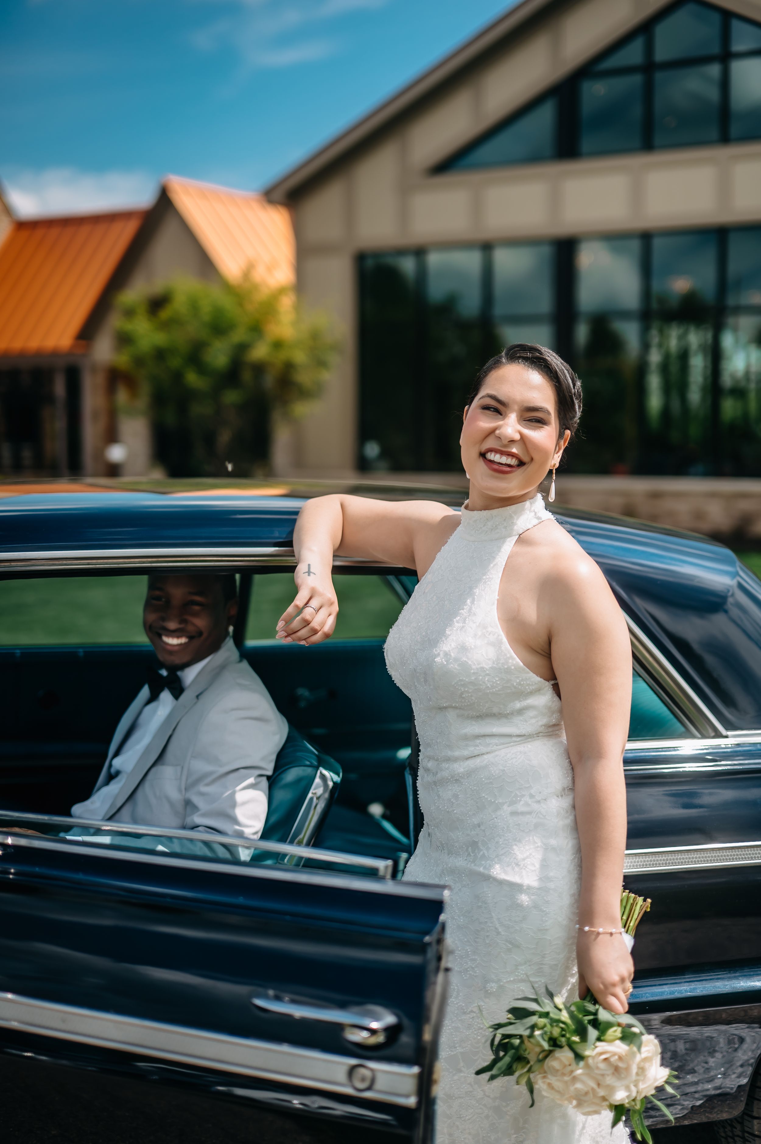 Bride in white halter gown holds bouquet beside vintage black car as groom smiles from the driver seat.