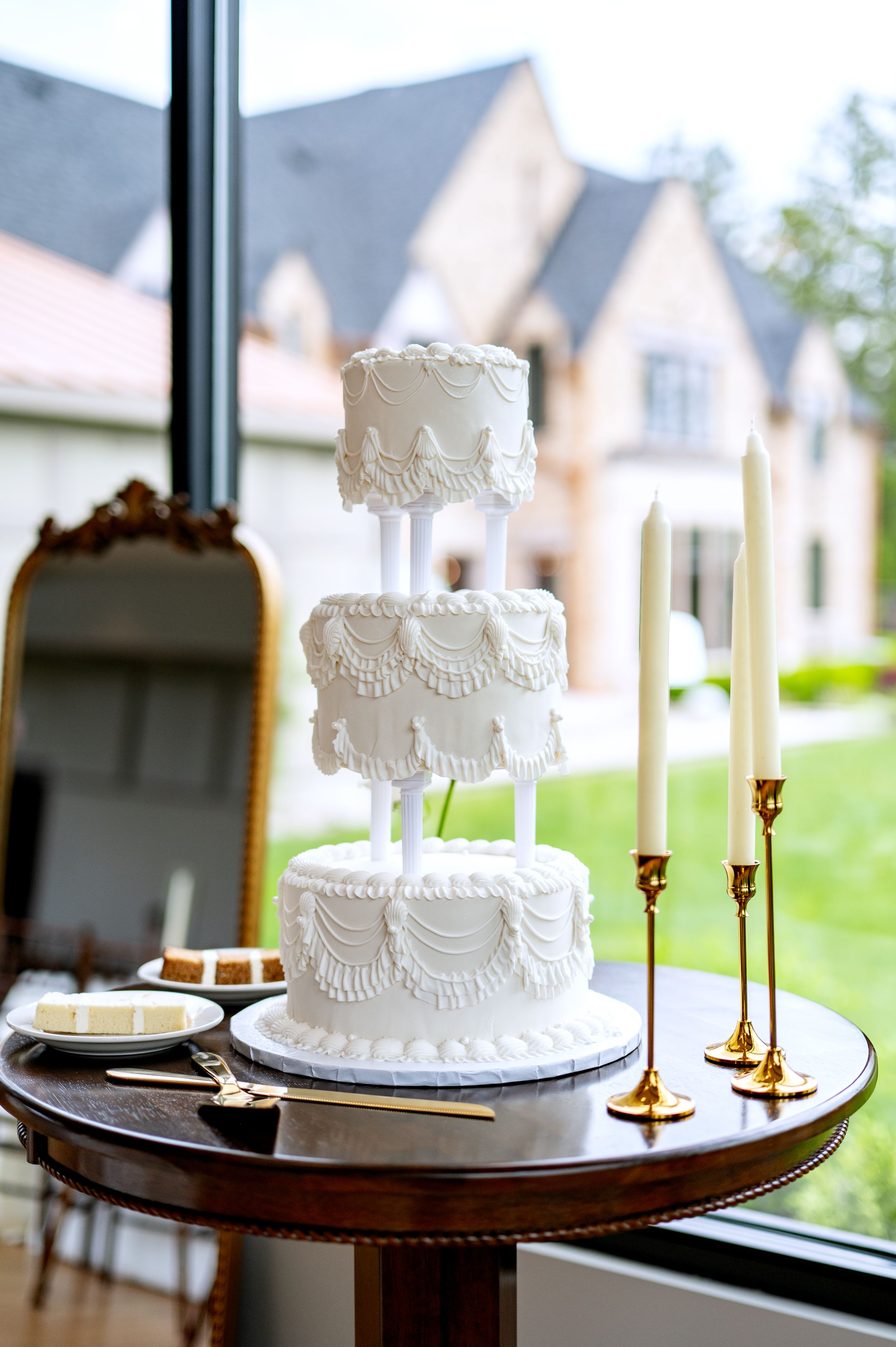 Elegant three-tier white wedding cake with intricate lace detailing, displayed on a wooden table with gold candlesticks.