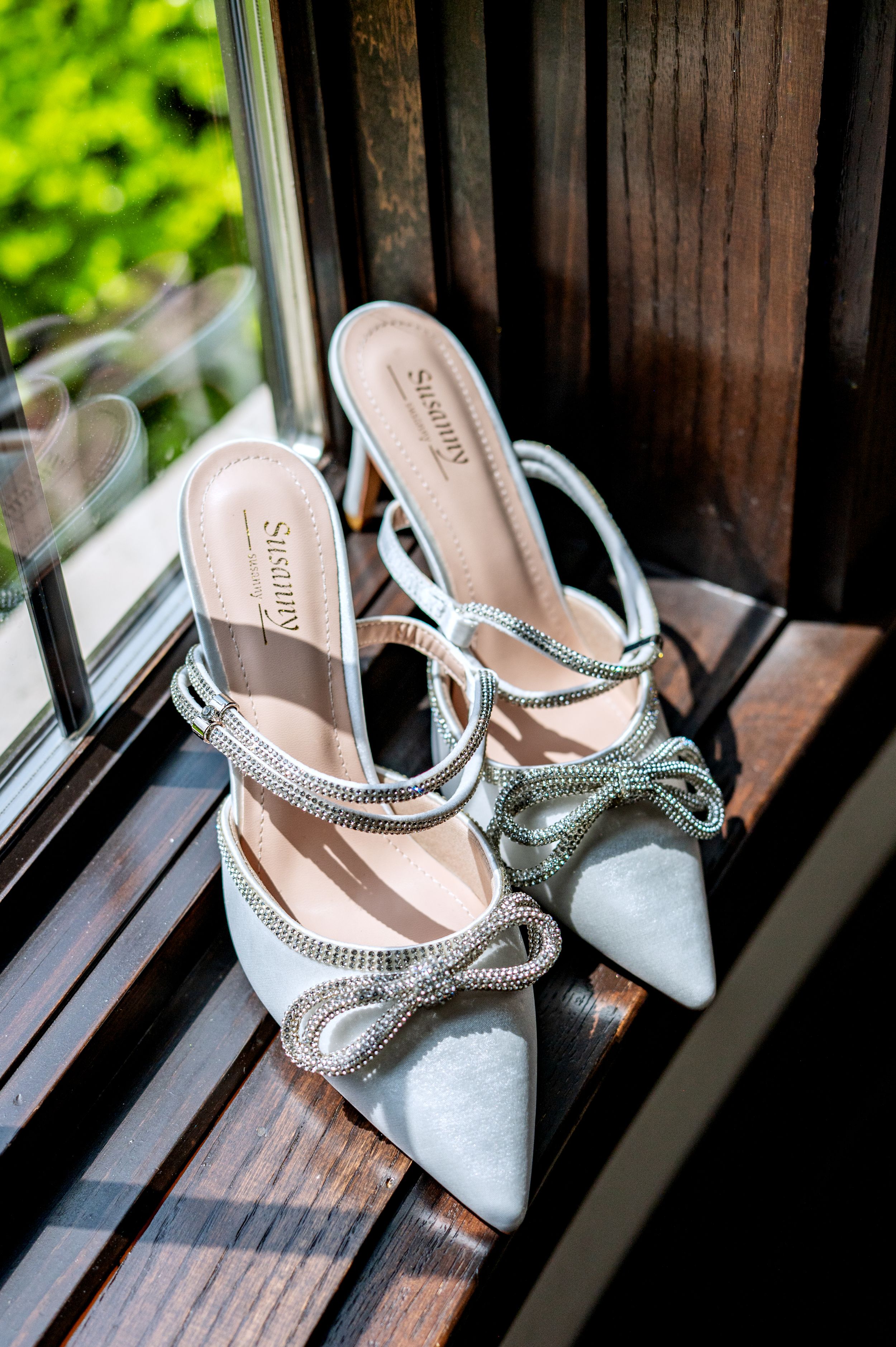 Elegant white satin bridal heels with rhinestone bow detail and ankle strap, displayed on a wooden windowsill.