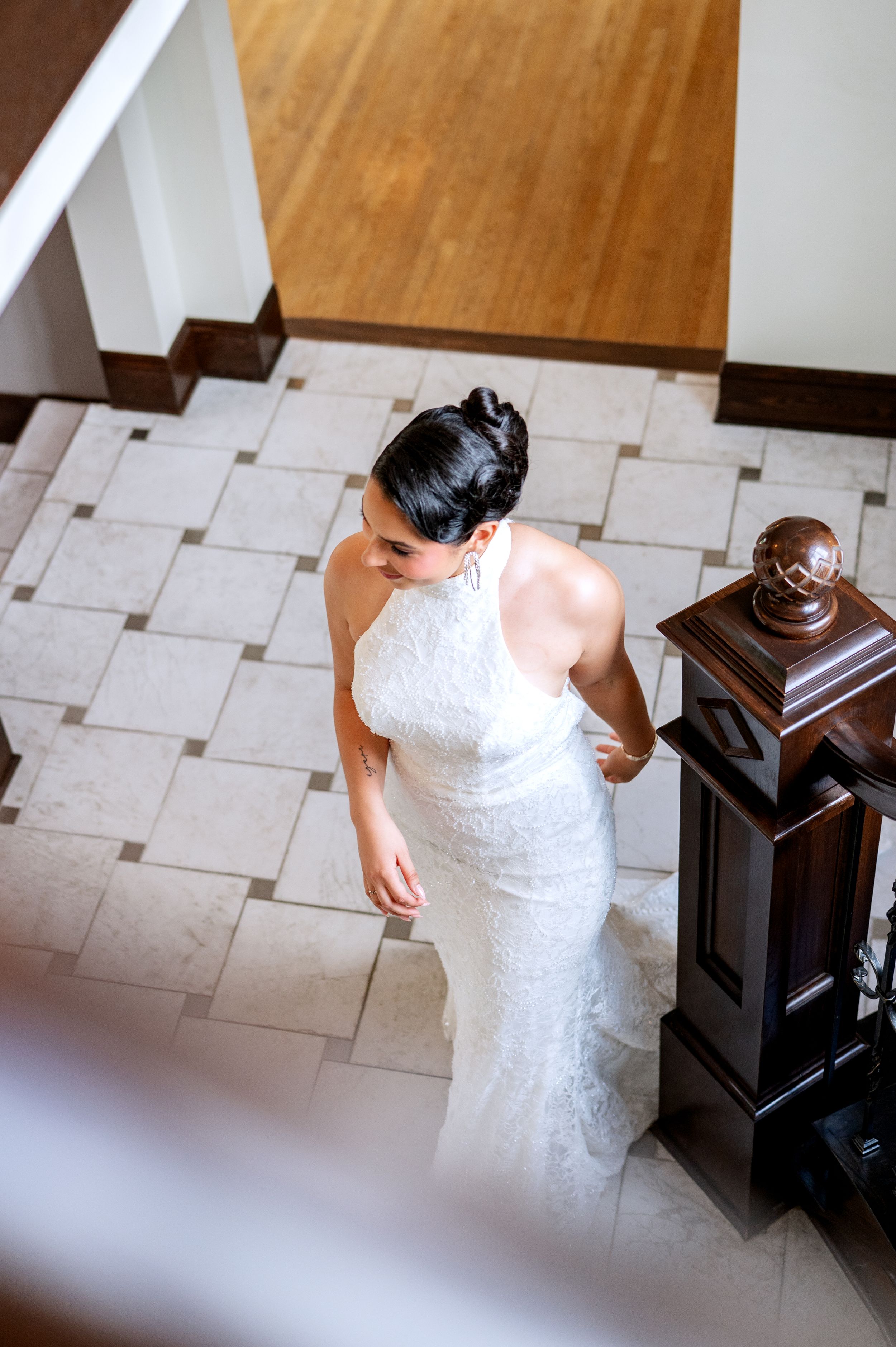 Aerial view of a bride in a white lace halter gown standing on a marble tile floor near a wooden newel post.