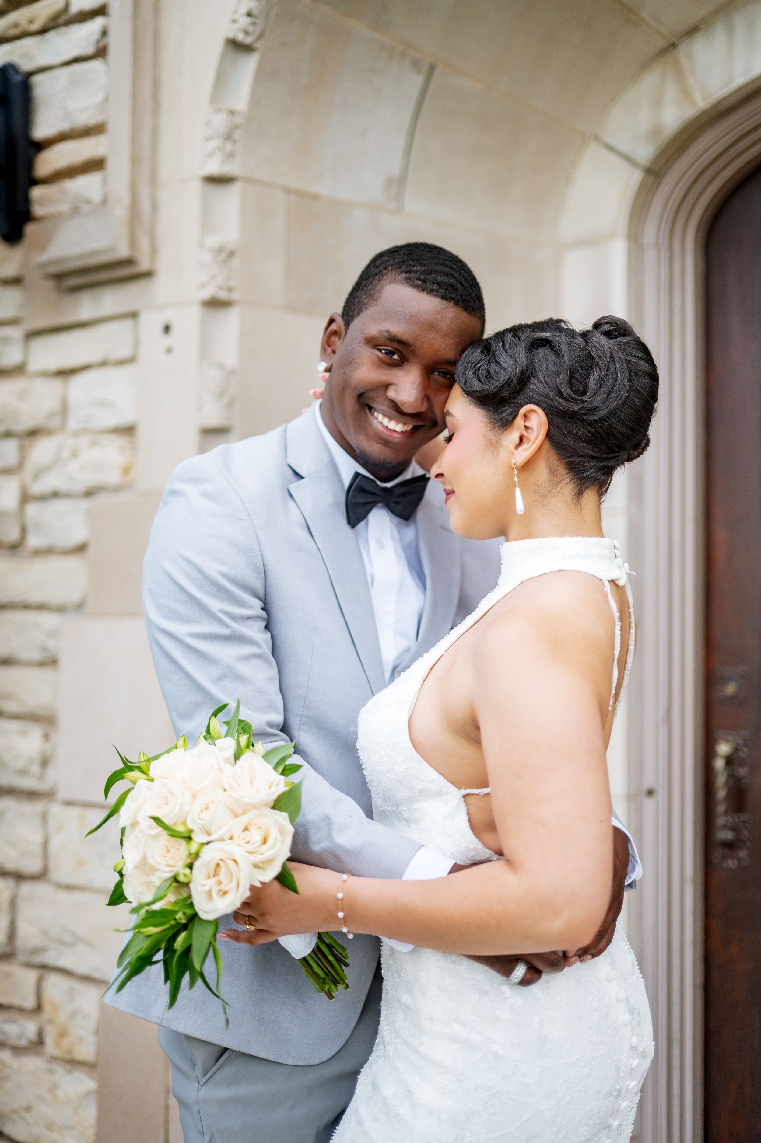 Smiling groom in gray suit and bow tie holds ivory rose bouquet while embracing bride in white halter gown outdoors.