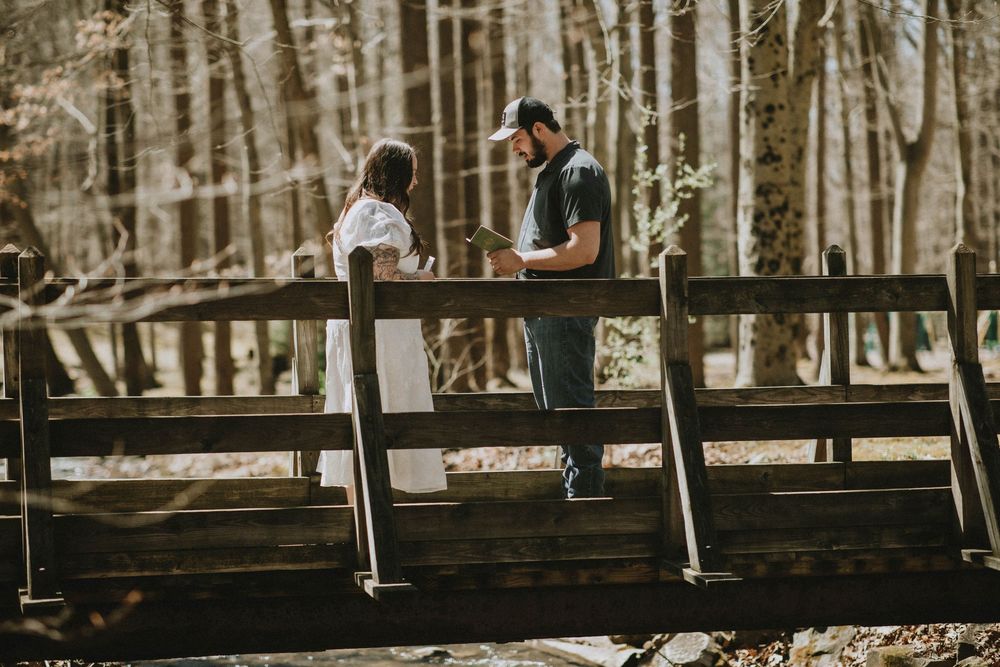 A couple stands together on a rustic wooden bridge surrounded by autumn forest trees in a romantic outdoor setting.