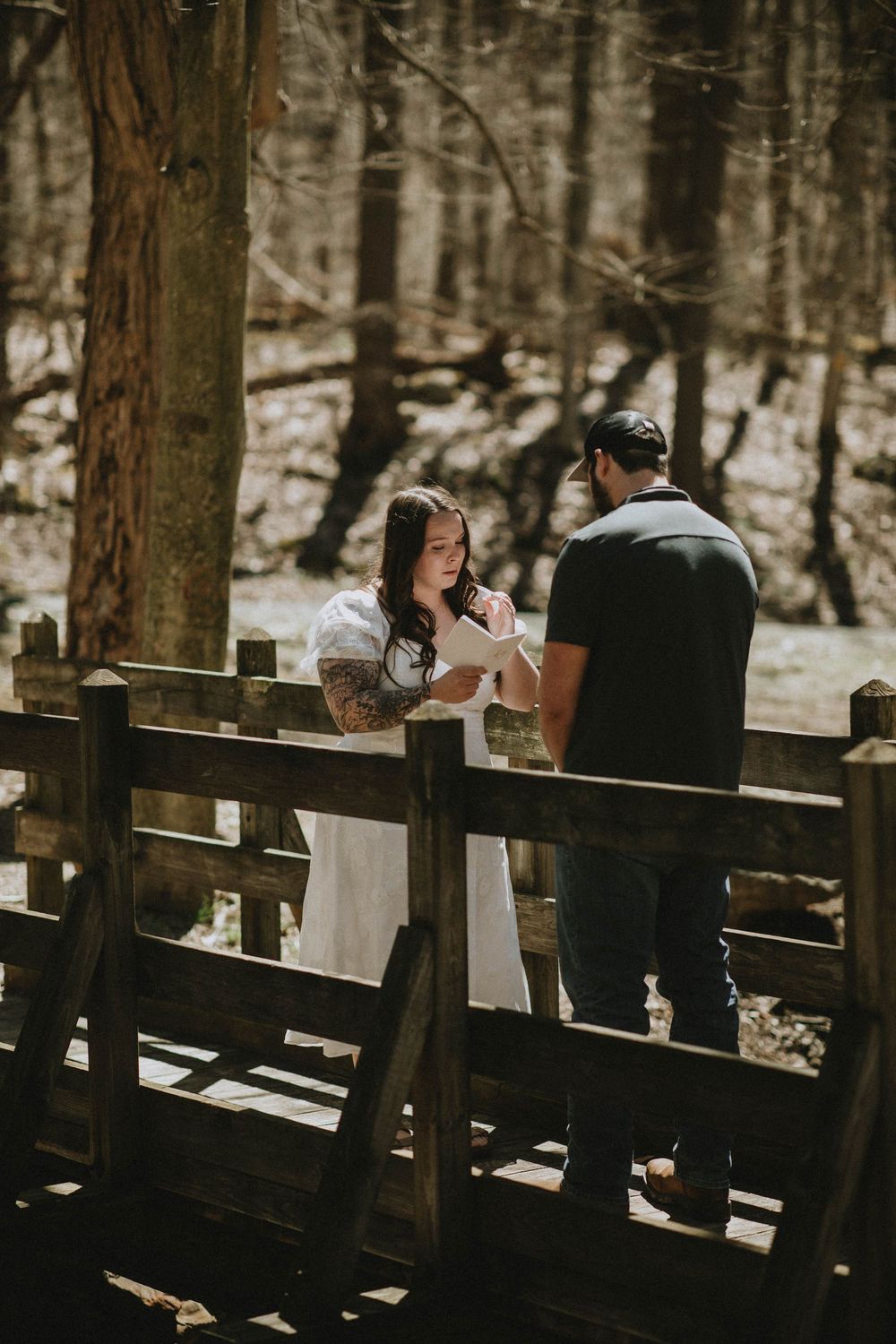 Couple sharing a romantic moment on a rustic wooden bridge in a forest, woman in white dress with tattoos.