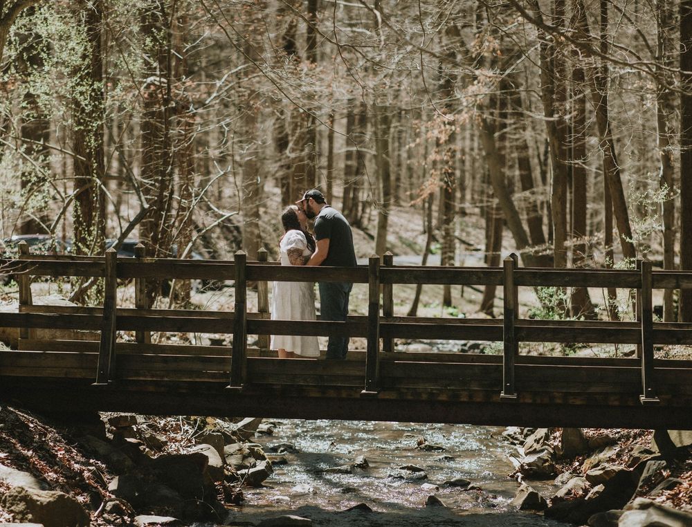 Couple sharing a romantic kiss on a wooden bridge over a stream in a lush forest setting. Blairsville PA photographer