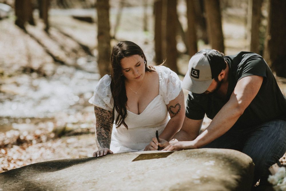 Bride in white lace dress and tattooed groom signing marriage license on rock in sunlit forest setting.