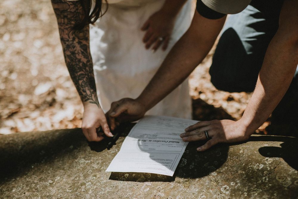 Bride and groom signing wedding marriage certificate on rustic wood surface, groom with tattooed arm.