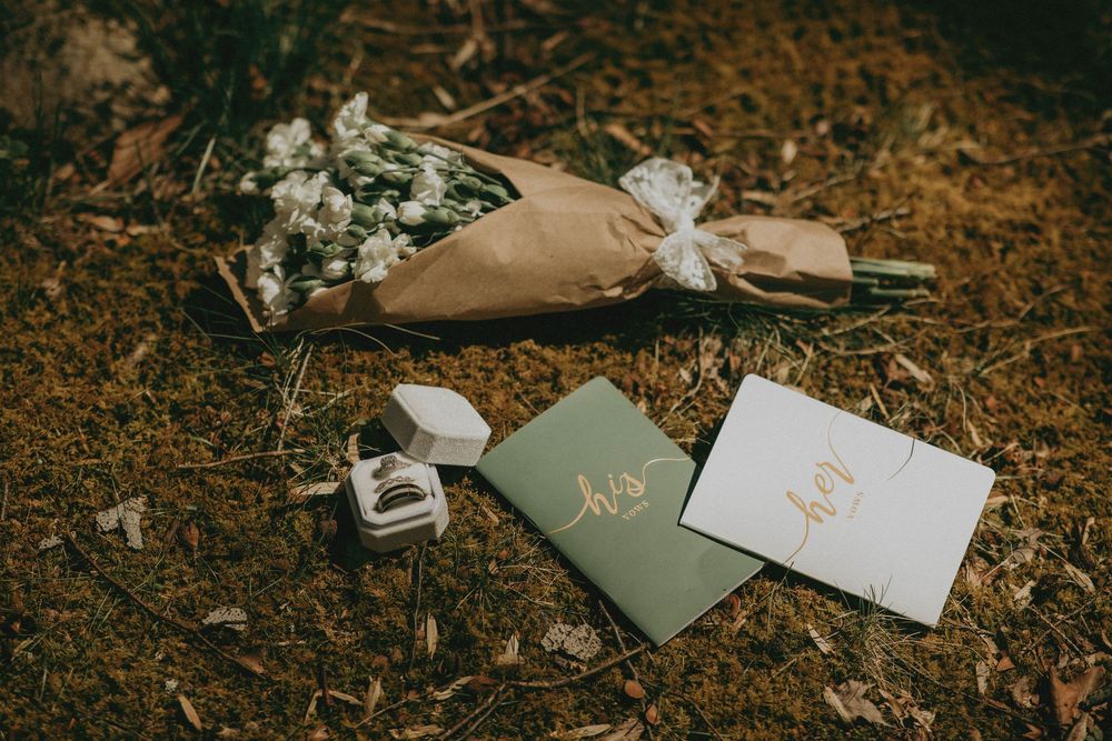 Wedding flat lay with floral bouquet, his and hers vow books, and ring box on mossy ground.
