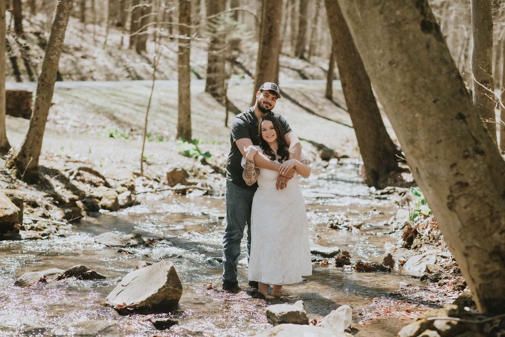 Couple embracing in a romantic woodland wedding photo beside a rocky stream surrounded by bare trees.