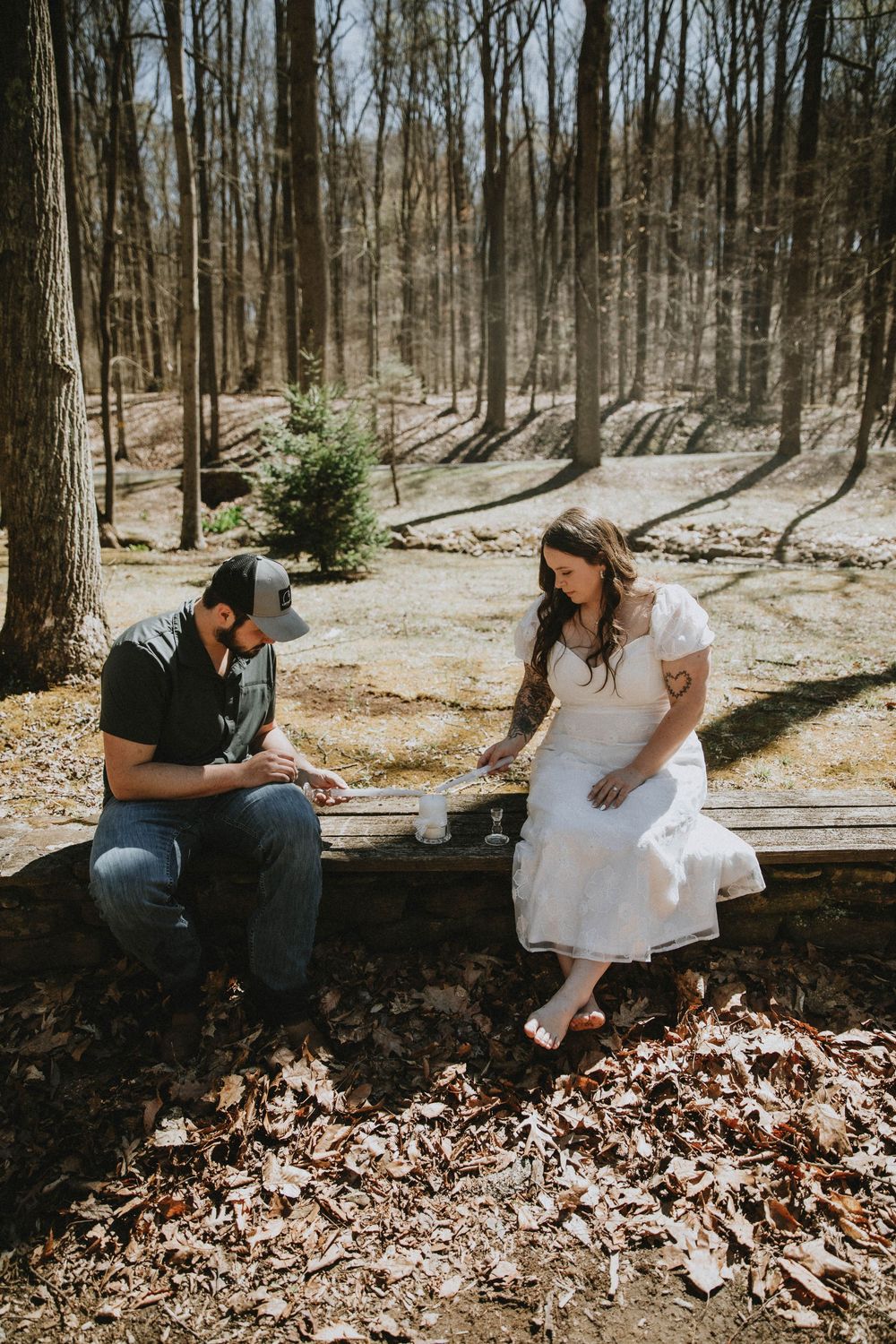 Couple in wedding attire sitting on a rustic bench in a sunlit forest, sharing a tender moment among autumn leaves.