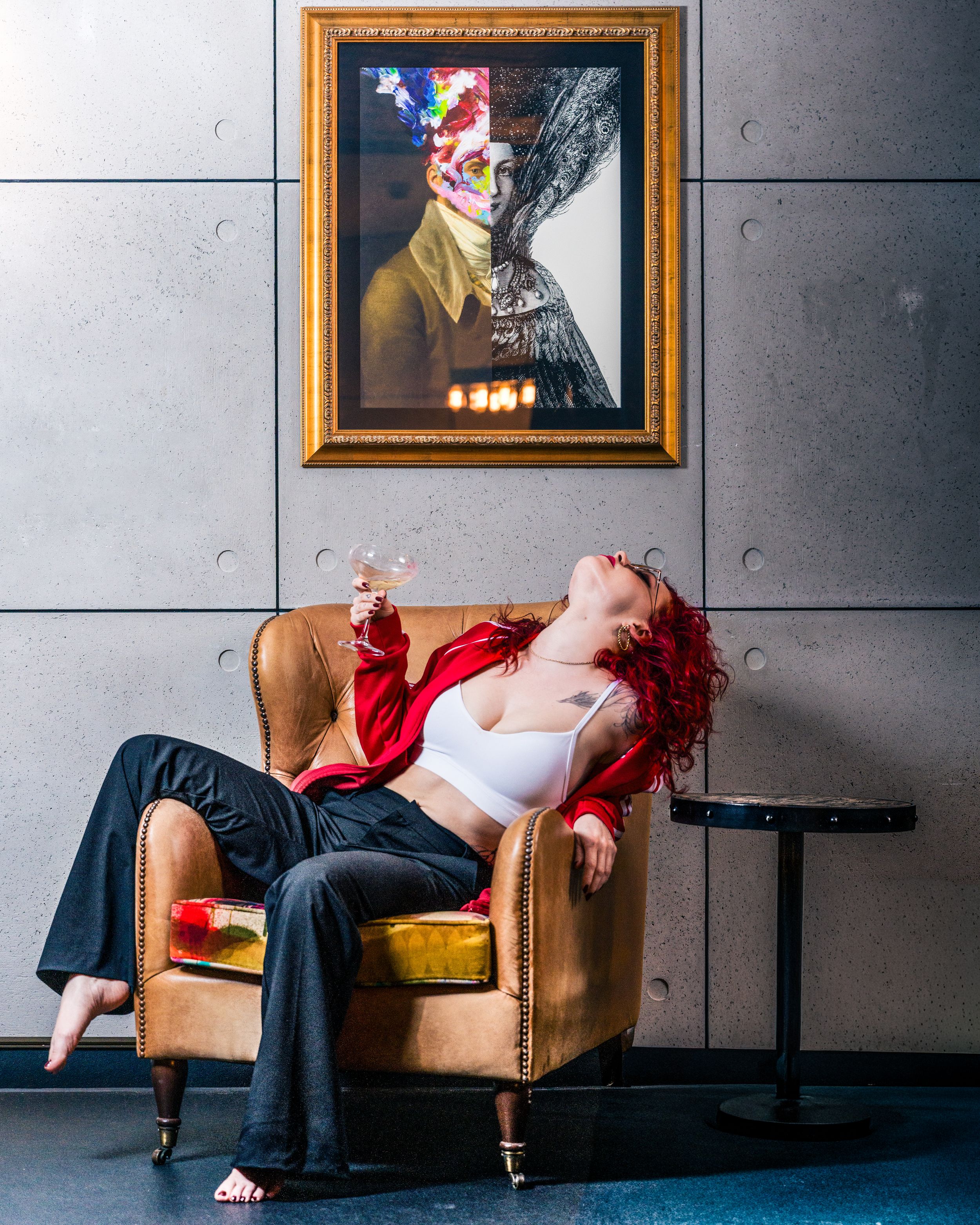 Editorial portrait of a model with red hair reclining in a gold armchair beside a framed painting