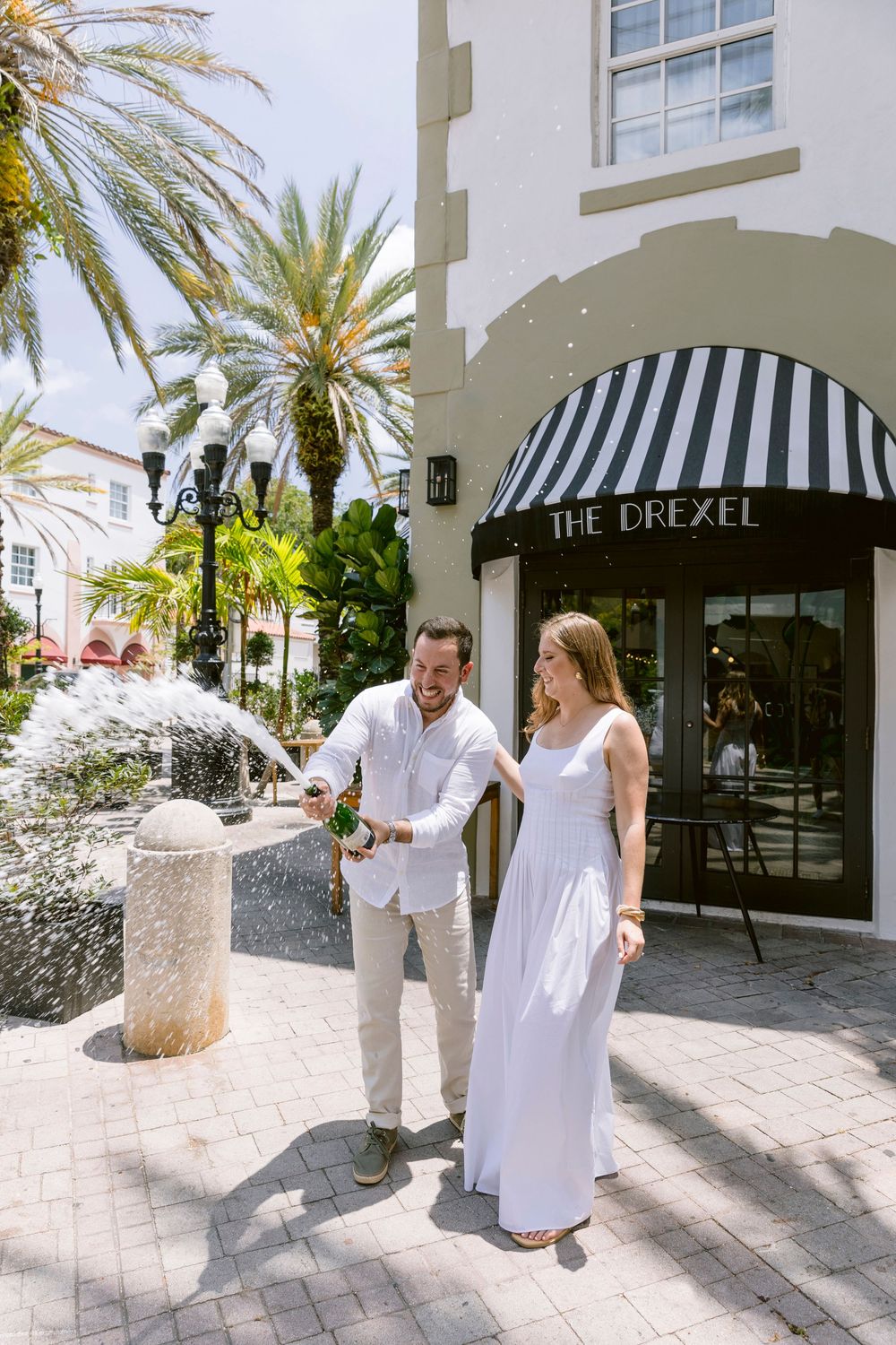 Couple in white outfits popping champagne outside The Drexel building with palm trees and fountain.