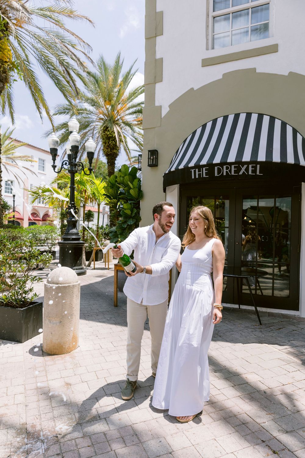 Couple in white celebrating with champagne outside The Drexel building, surrounded by palm trees on a sunny day.