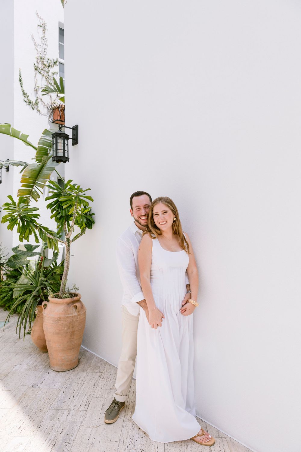Happy couple in white outfits posing against a white wall with tropical plants in a sunny outdoor setting.