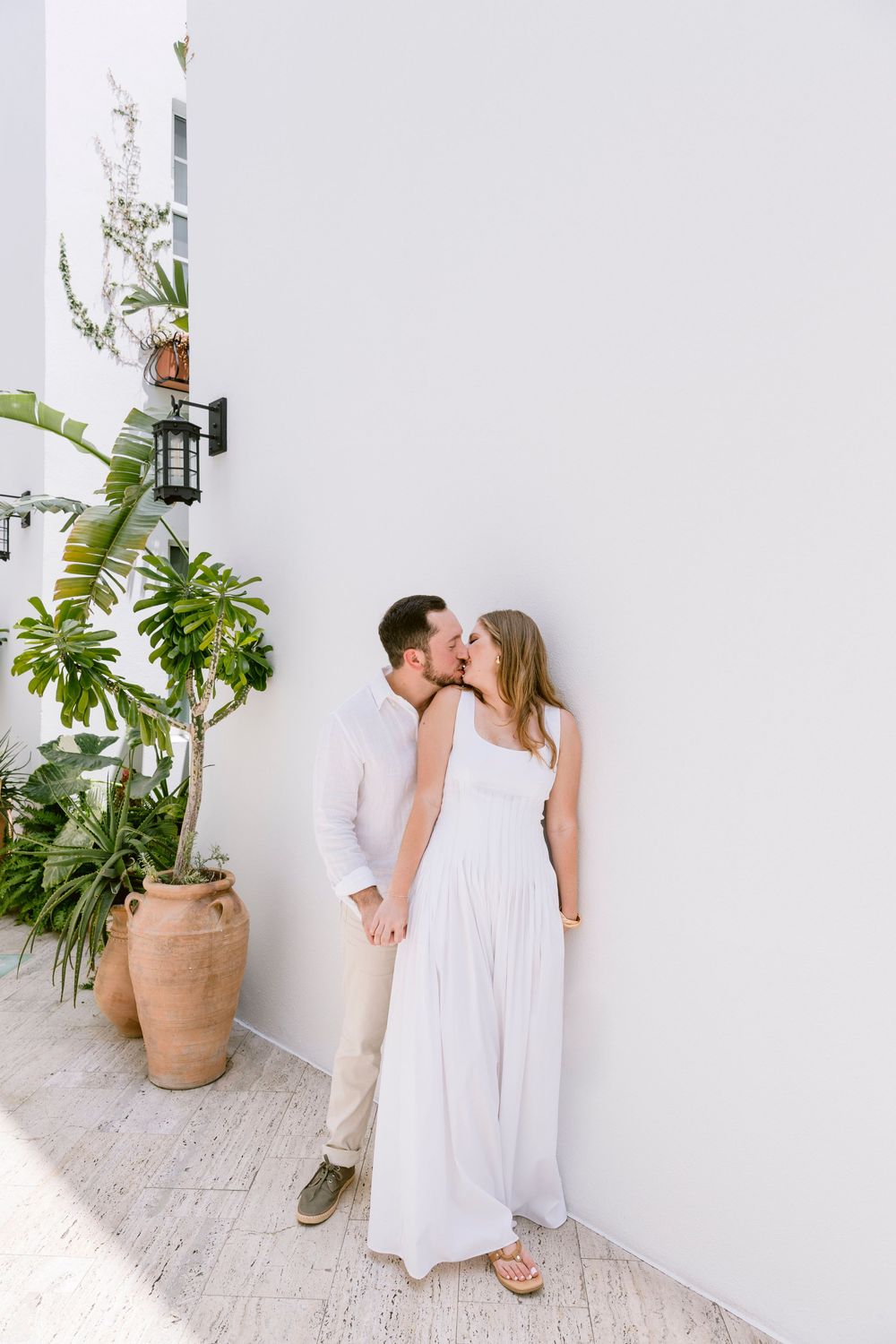A couple shares a romantic kiss against a white wall, surrounded by lush tropical plants in terracotta pots.