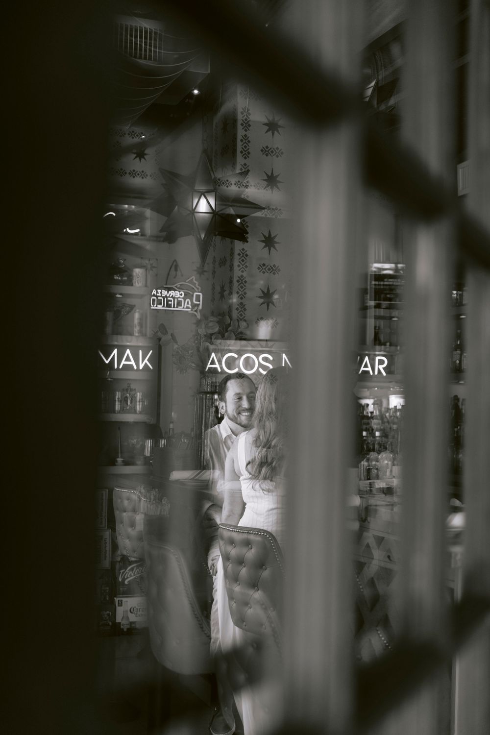 Black and white photo of a smiling woman seen through a window with reflections of neon signs reading MAK TACOS AR.