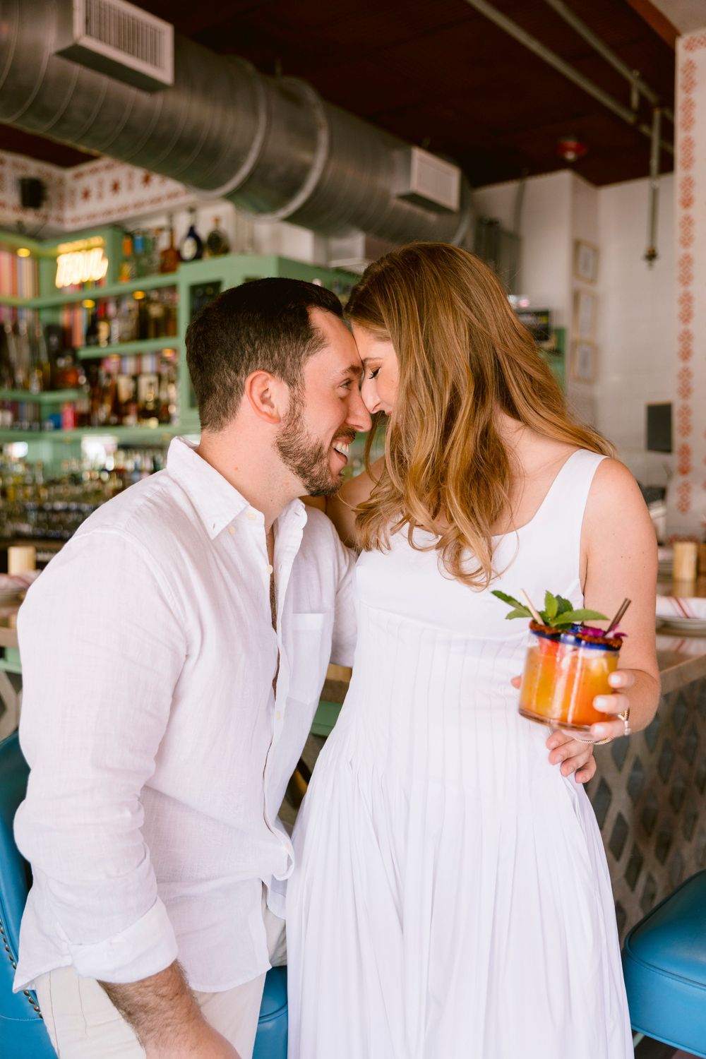A couple in white outfits share a romantic moment at a bar, woman holding a colorful cocktail garnished with mint.