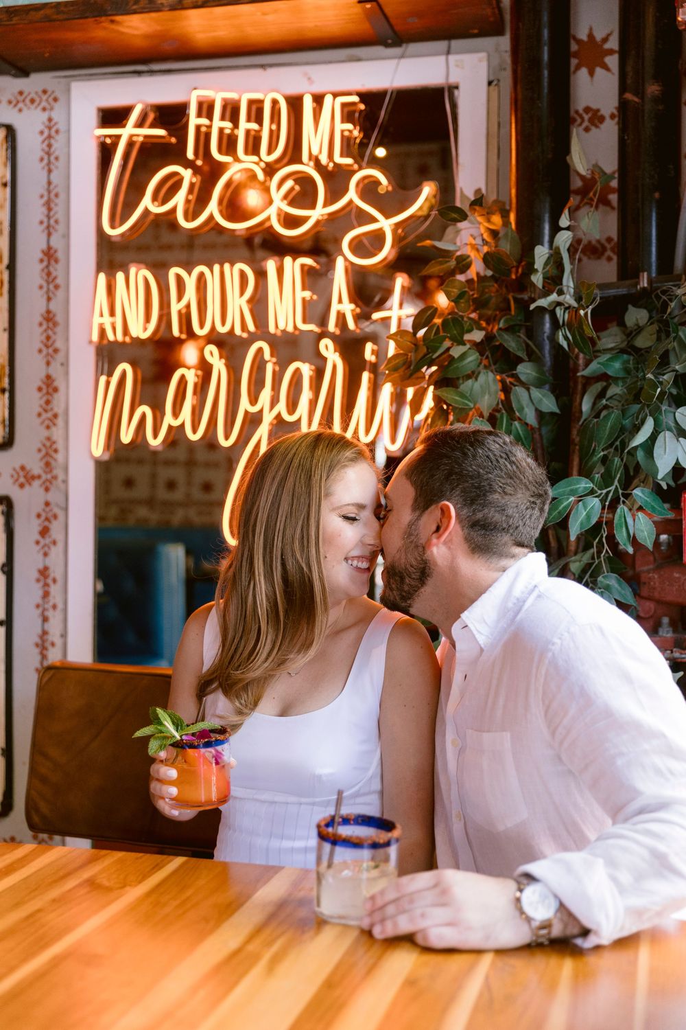 Couple sharing a romantic moment at a Mexican restaurant bar, with a glowing taco and margarita neon sign behind them.