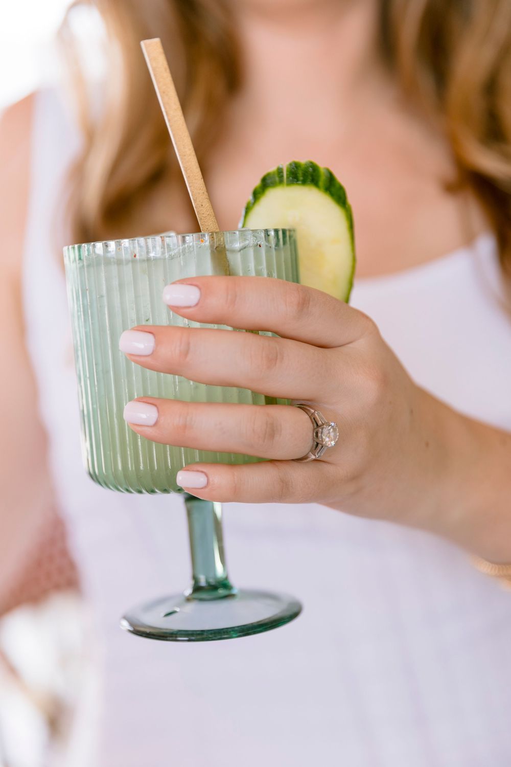 Bride holding a green ribbed cocktail glass with cucumber garnish, showing engagement ring.