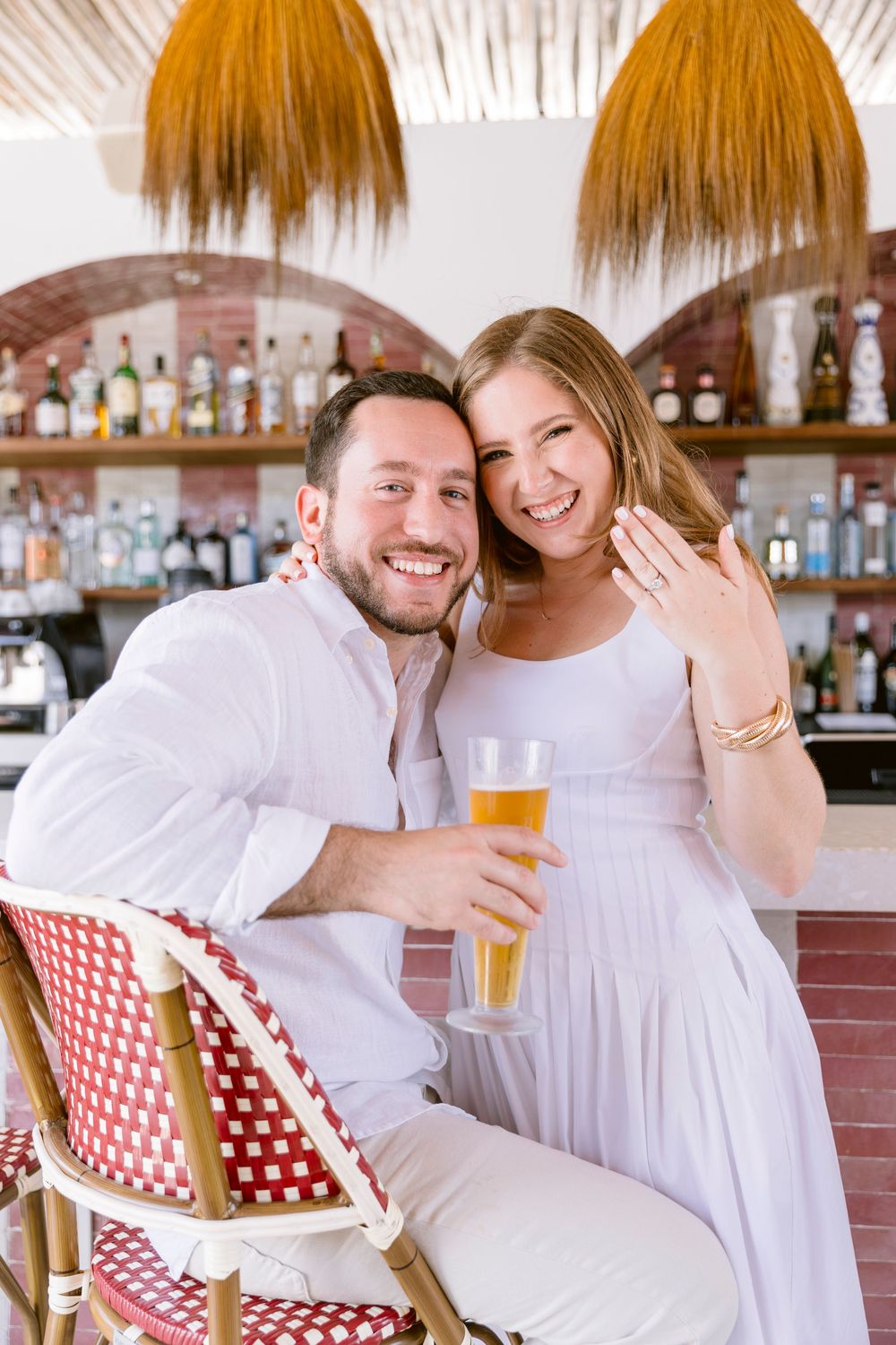 Engaged couple smiling at bar, man holding beer, woman showing diamond ring.