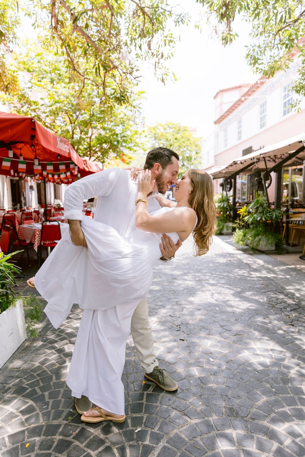 Couple in white attire sharing a romantic dip kiss on a cobblestone street surrounded by lush trees.