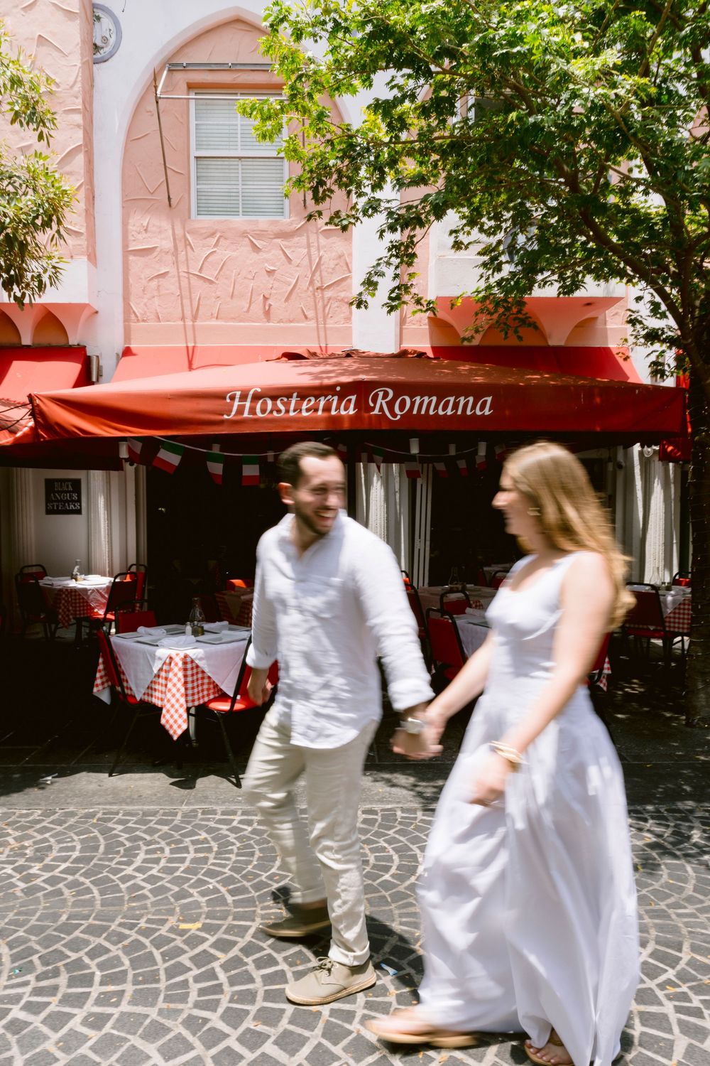 Couple holding hands walking past Hosteria Romana restaurant with red awning on cobblestone street in Rome.