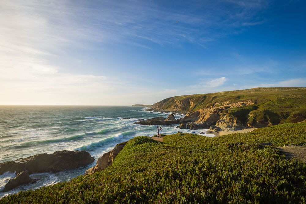 Scenic coastal landscape with rolling green hills, rocky shoreline, and two people standing at sunset.