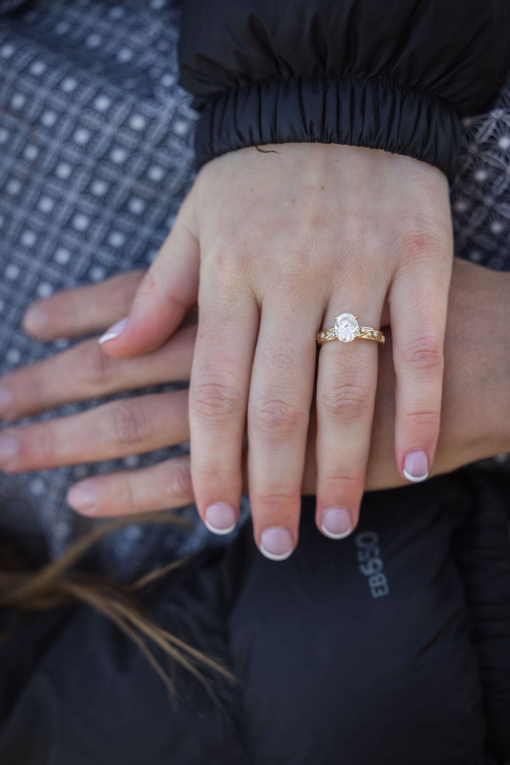 Close-up of a woman's hand displaying a gold engagement ring with oval diamond solitaire, resting on a man's hand.
