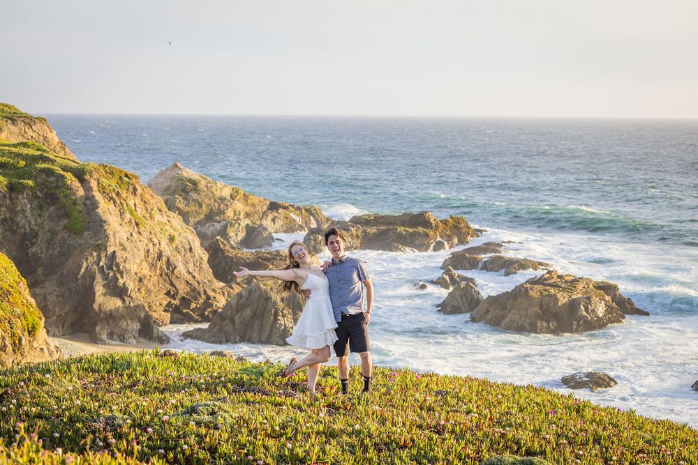Happy couple poses on a scenic coastal bluff with crashing ocean waves in the background.