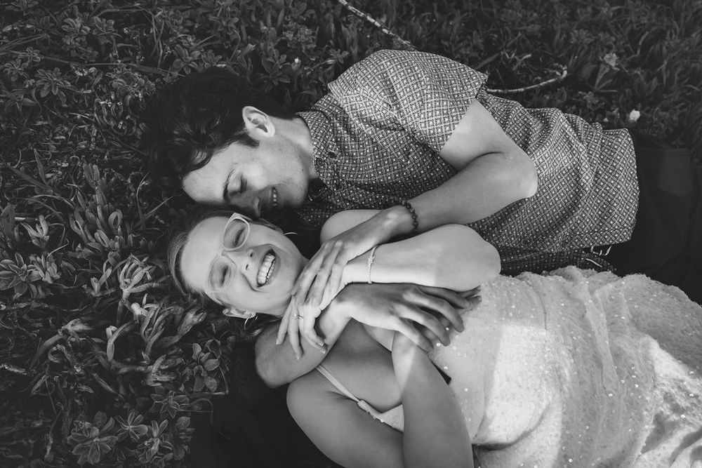 Black and white photo of a smiling couple lying on grass, embracing tenderly in a romantic outdoor setting.