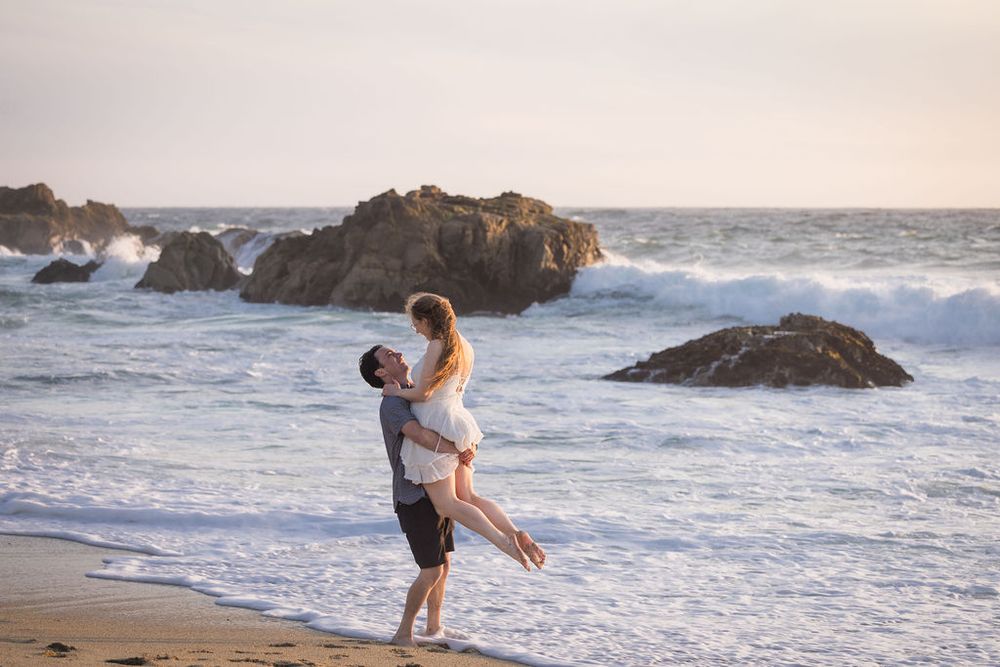 Romantic couple embracing on a scenic beach at sunset with waves crashing on rocks in the background.