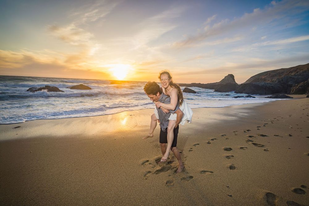 Couple sharing a piggyback ride on a sandy beach at golden sunset with waves and rocky coastline in background.