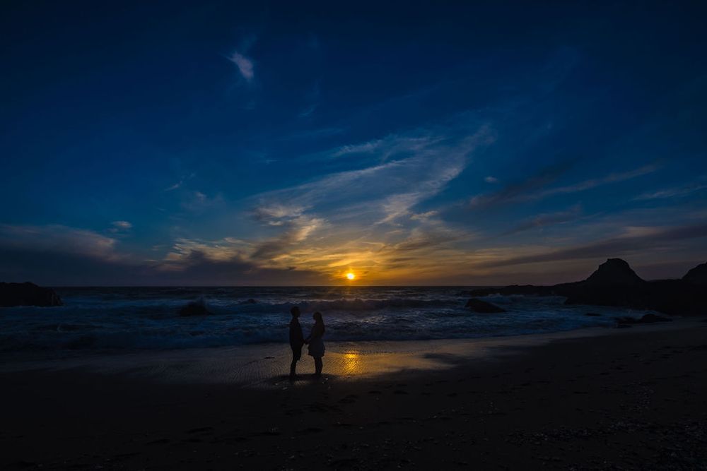 Silhouette of couple standing on beach at sunset with golden horizon, ocean waves, and dramatic blue sky.