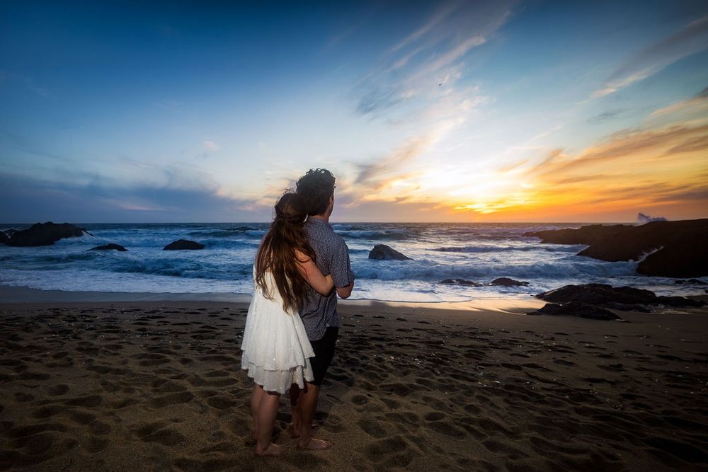 Romantic couple watching a stunning sunset together on a rocky beach, woman in white dress.