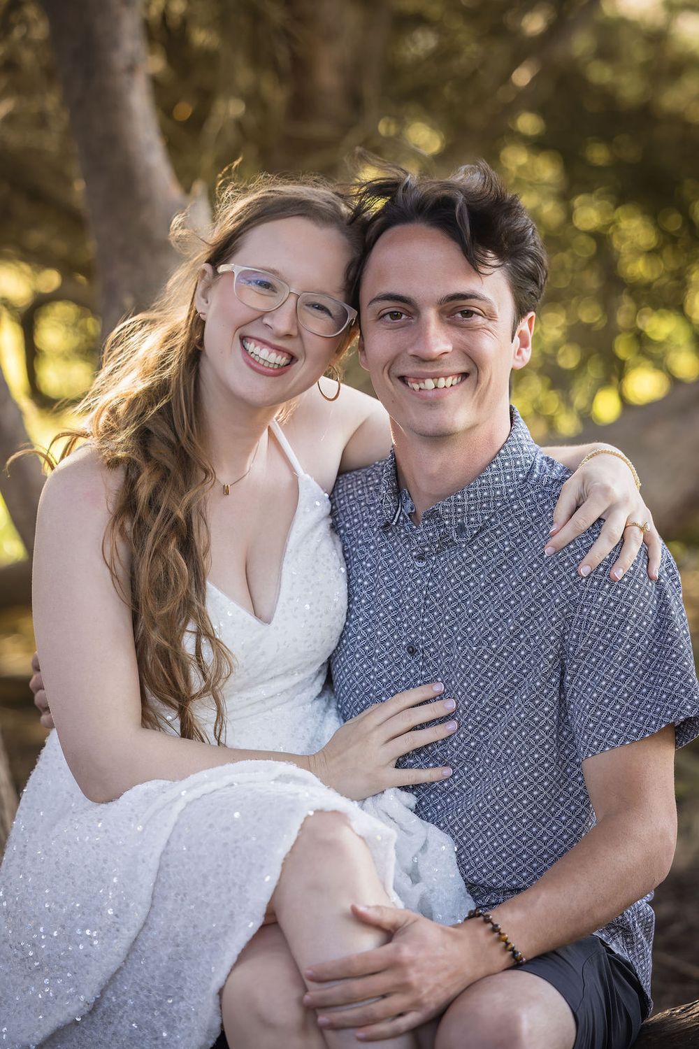 Smiling couple embracing outdoors, woman in white dress with glasses sitting on man's lap among trees.