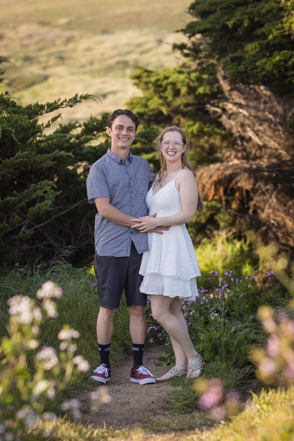 A smiling couple poses on a scenic coastal trail surrounded by wildflowers and trees during golden hour.