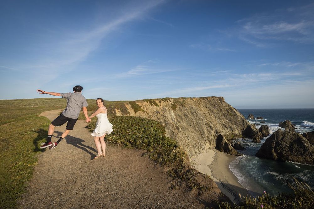 Couple holding hands on coastal cliffside trail with dramatic ocean and rock formations in background.