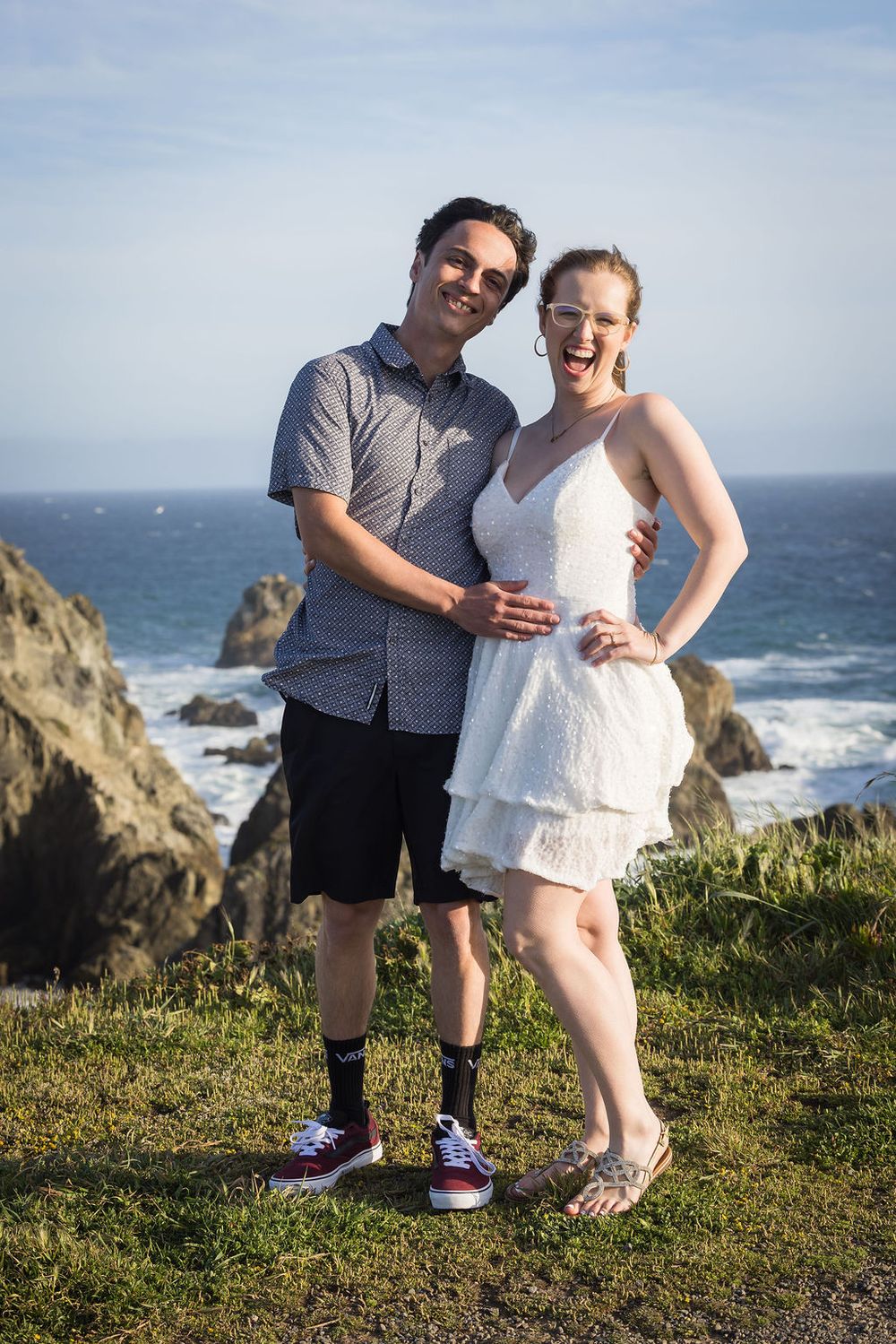 Smiling couple poses together on a coastal cliff with ocean waves and rocks behind them.