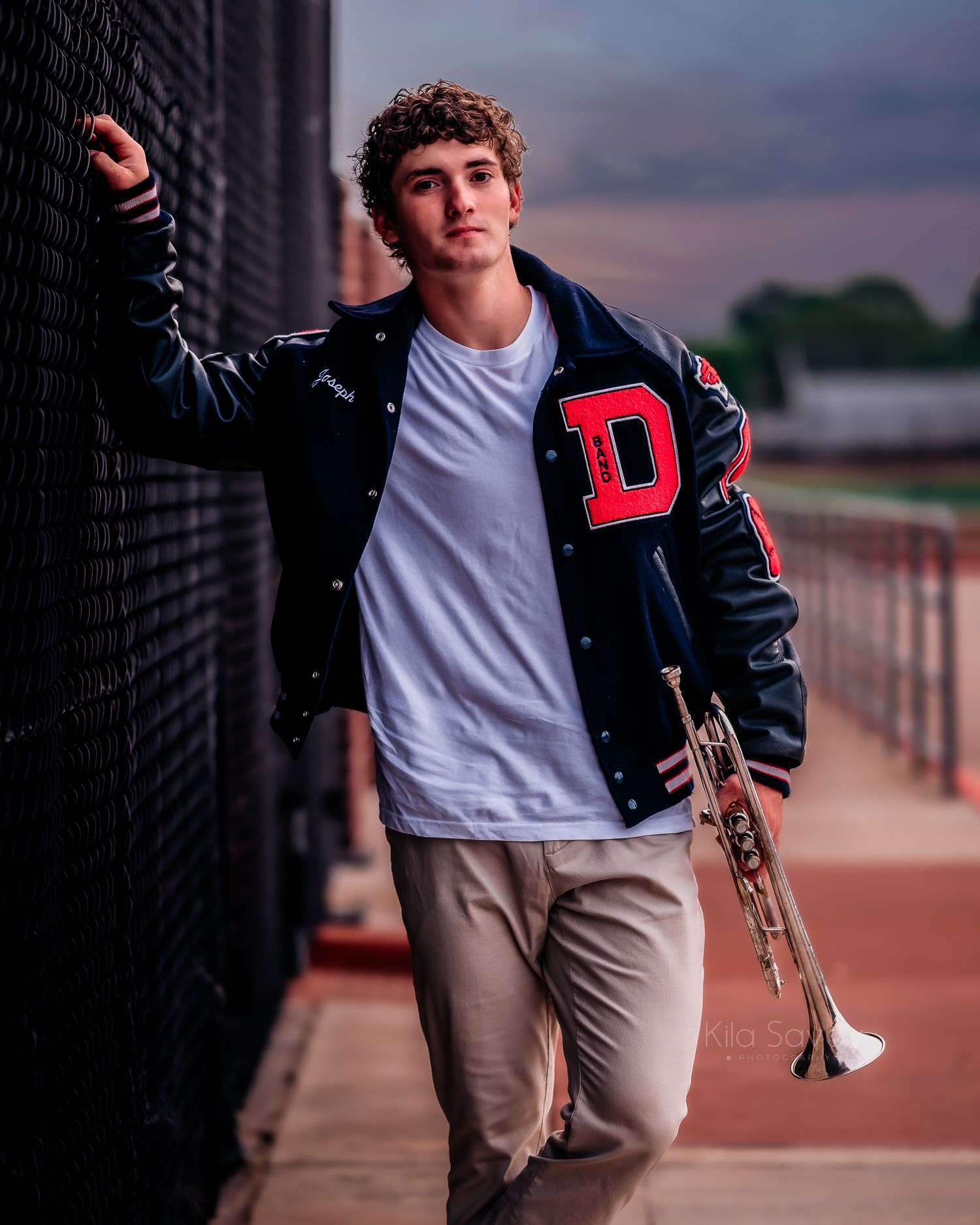 Young man in a varsity jacket holding a trumpet leans against a fence at a stadium for senior portraits.