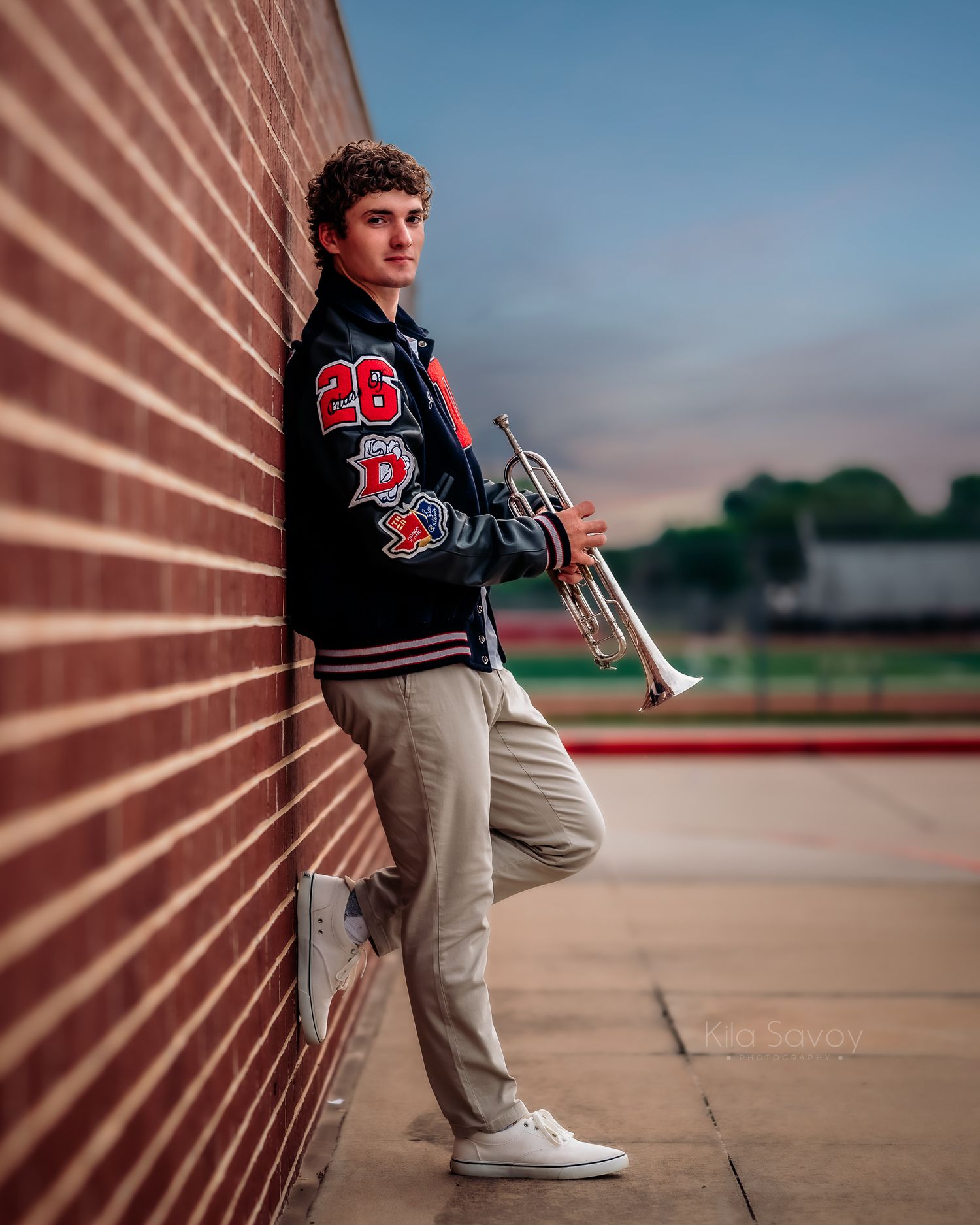 Teen boy in varsity jacket holding trumpet leans against brick wall outdoors at dusk for senior portrait photo.