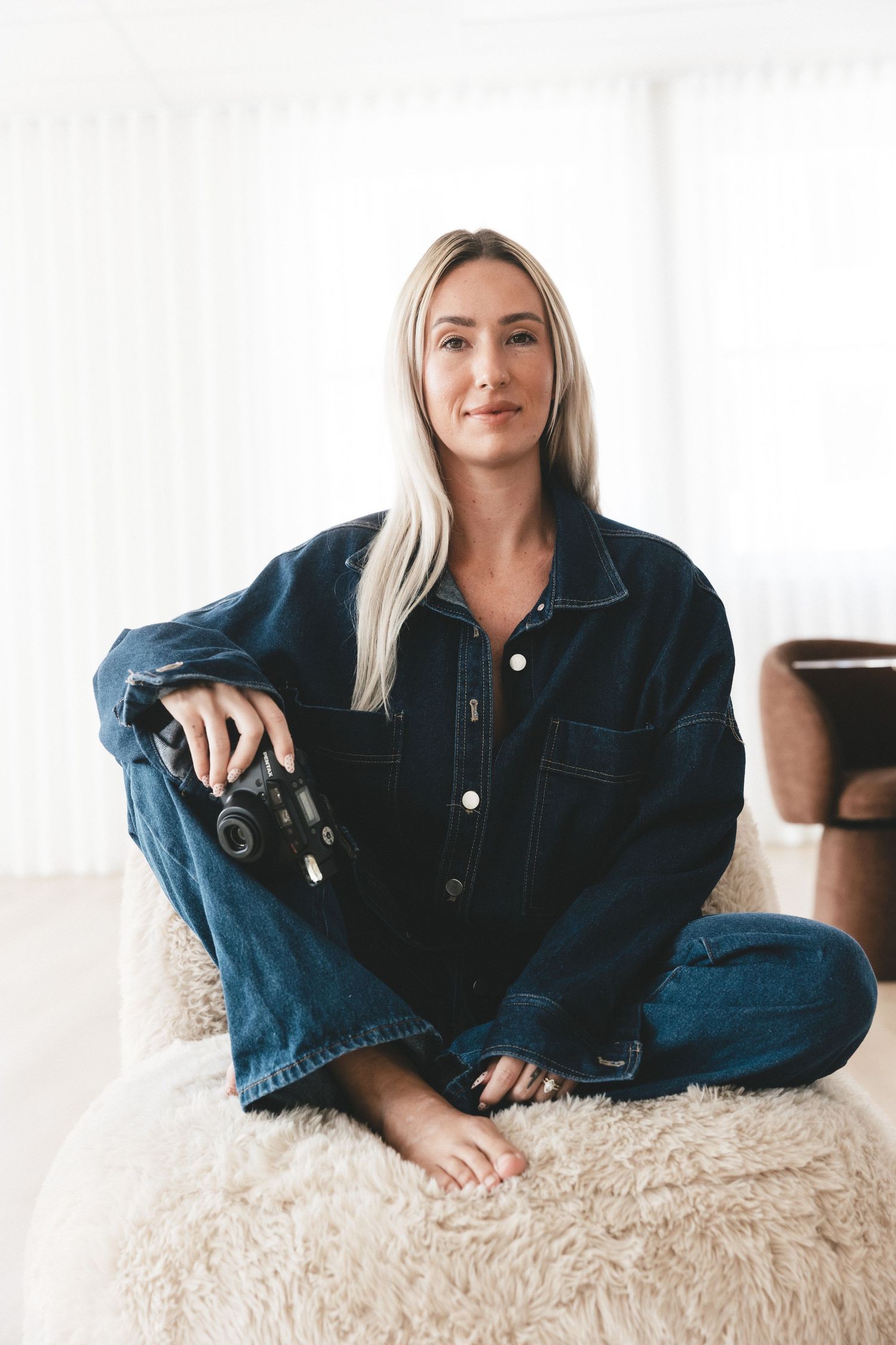 Blonde woman in denim jacket and jeans sitting cross-legged on fluffy ottoman holding a camera indoors.