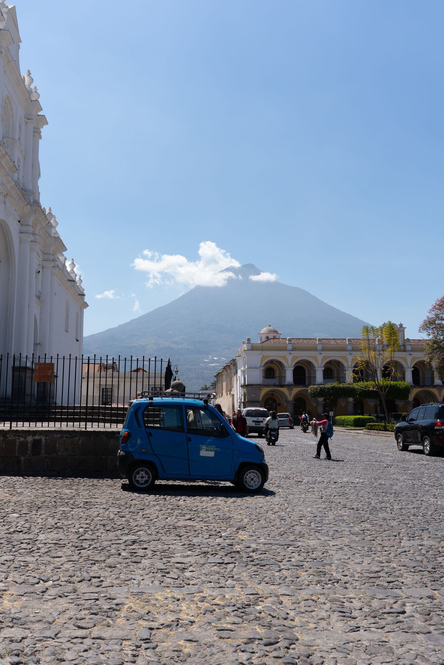 A small blue car parked in a cobblestone plaza with Agua Volcano rising dramatically in the background in Antigua, Guatemala.