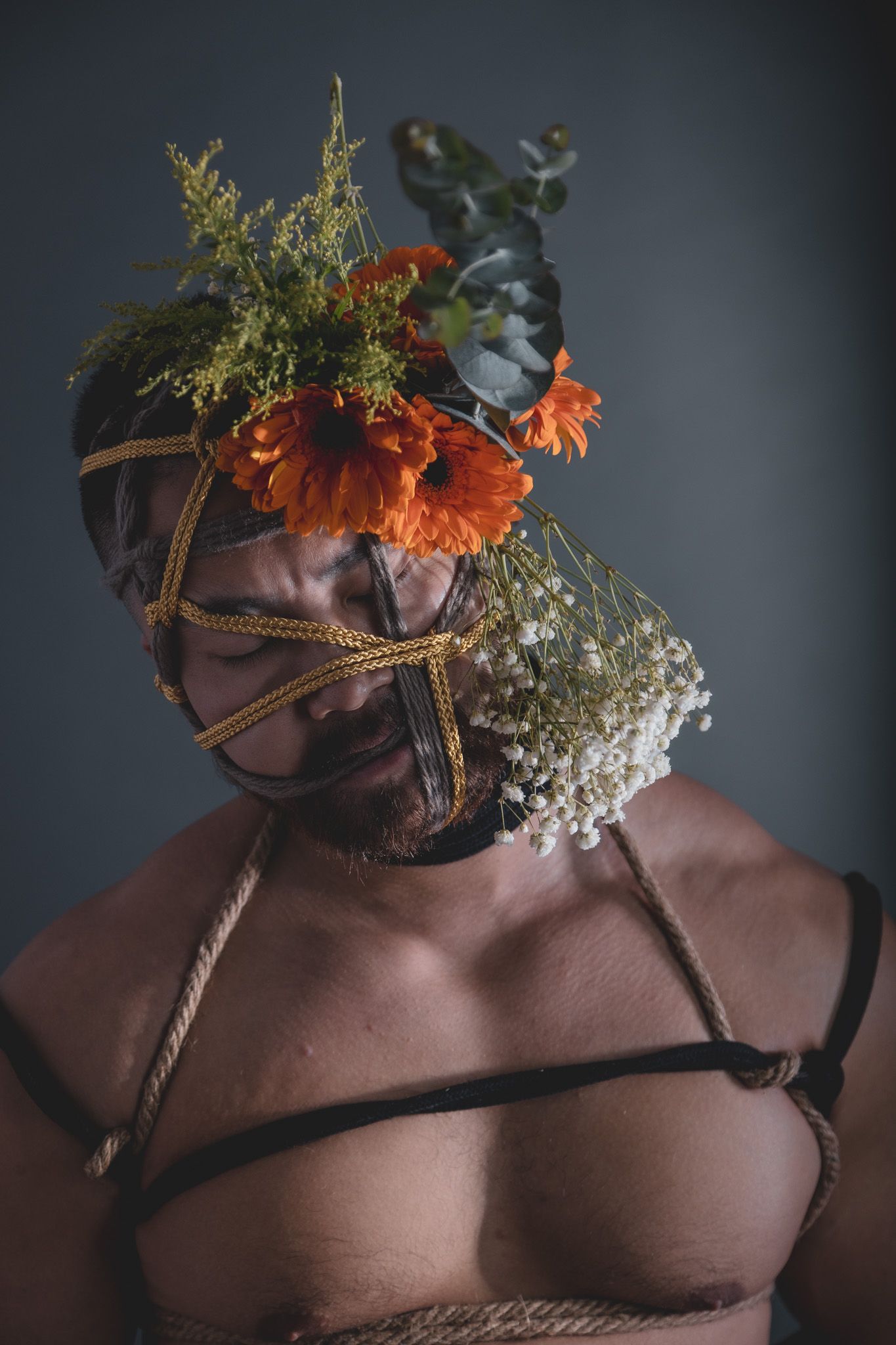 Shirtless man with face bound in rope and orange flowers arranged as a floral mask, studio portrait.