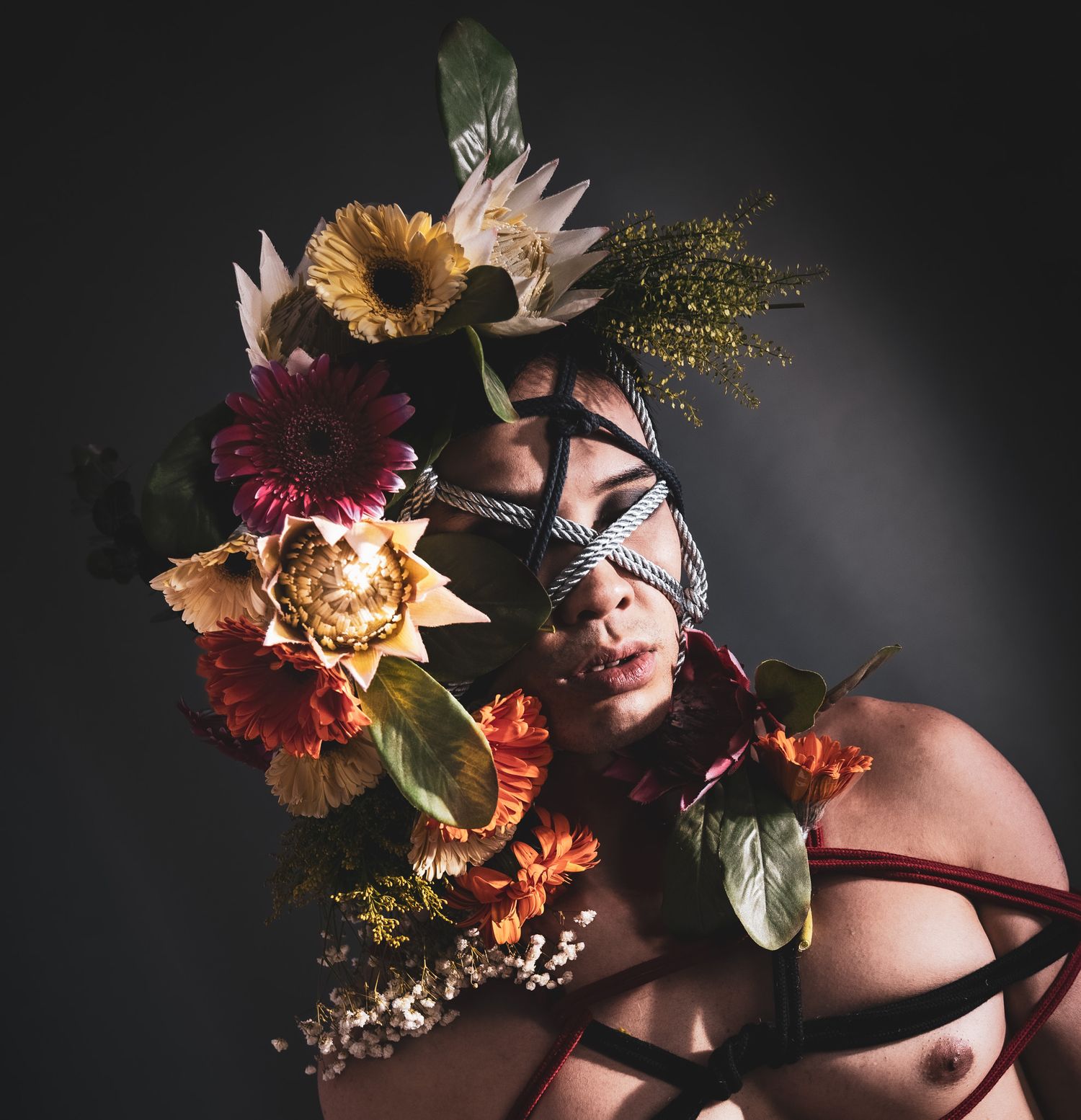 Close-up artistic portrait of a person with rope face binding and vibrant floral arrangement on head.