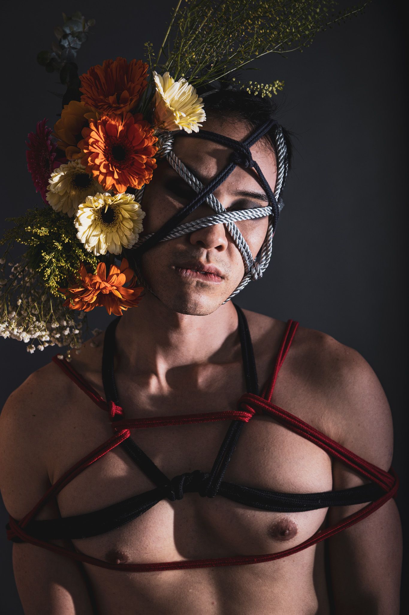 Studio portrait of a person with rope face binding, floral crown, and red and black body harness.