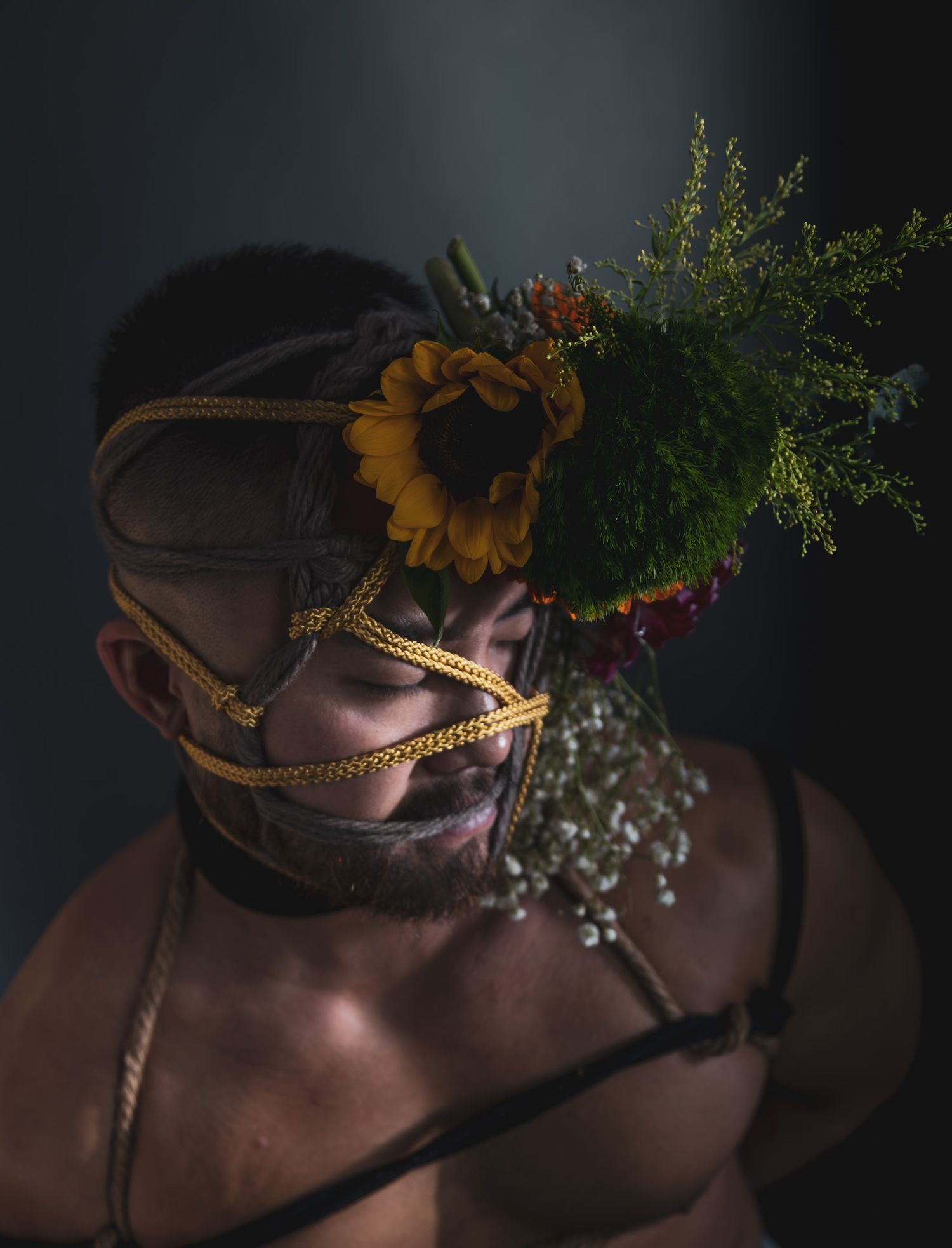 Close-up of person with yellow braided rope face bondage adorned with sunflower and green foliage.
