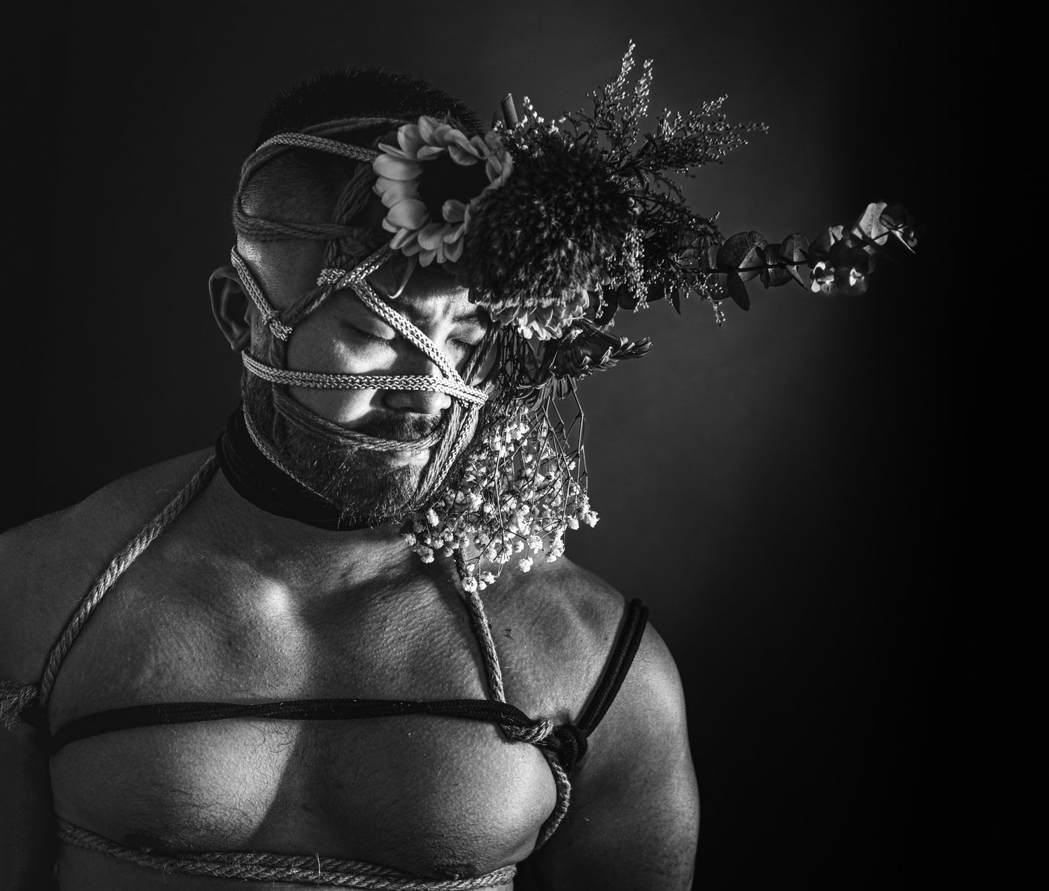 Black and white portrait of a person wearing a leather mask adorned with dried flowers and a harness.