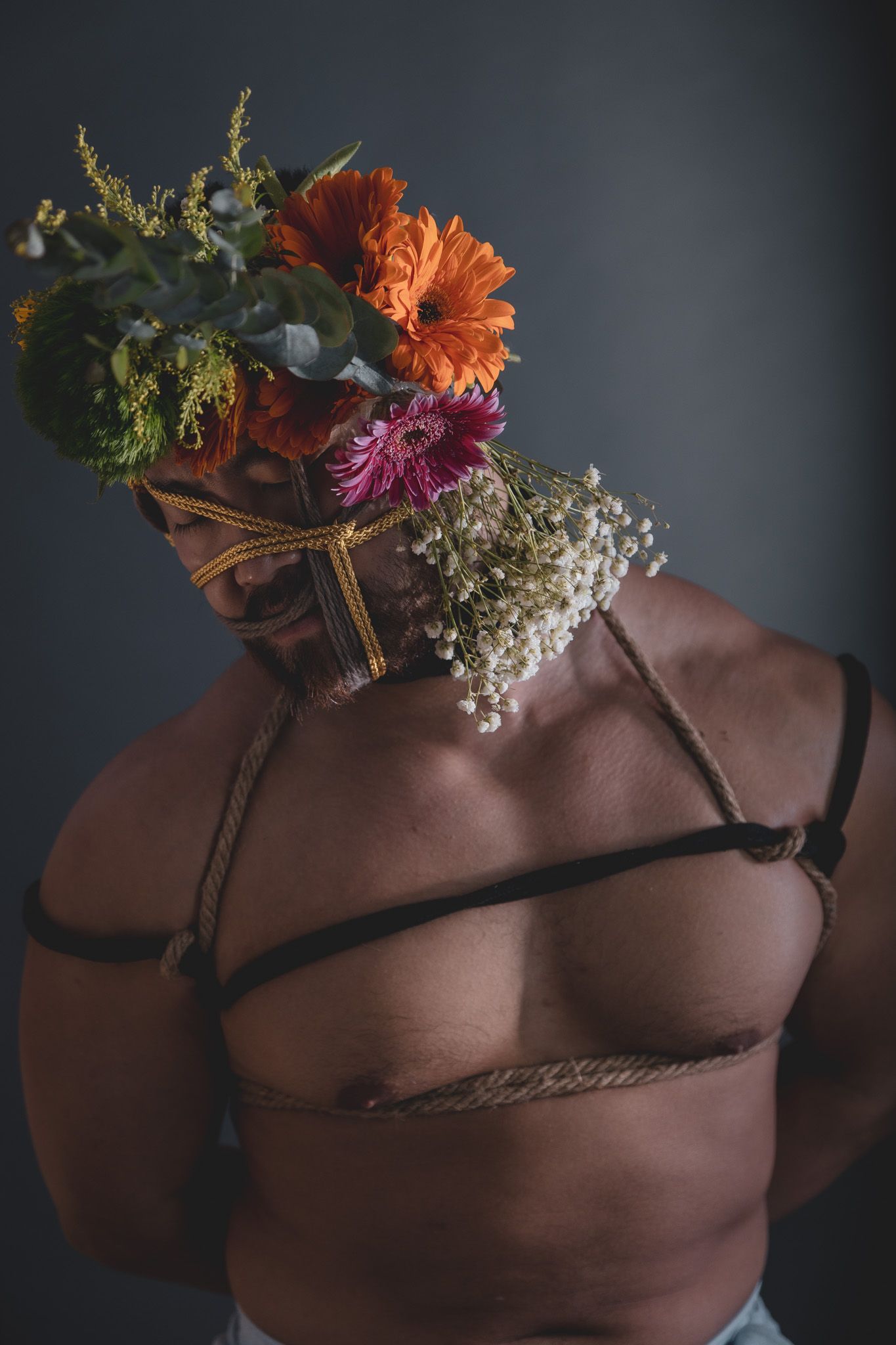 Artistic portrait of a muscular man with rope harness and colorful floral arrangement covering his face.