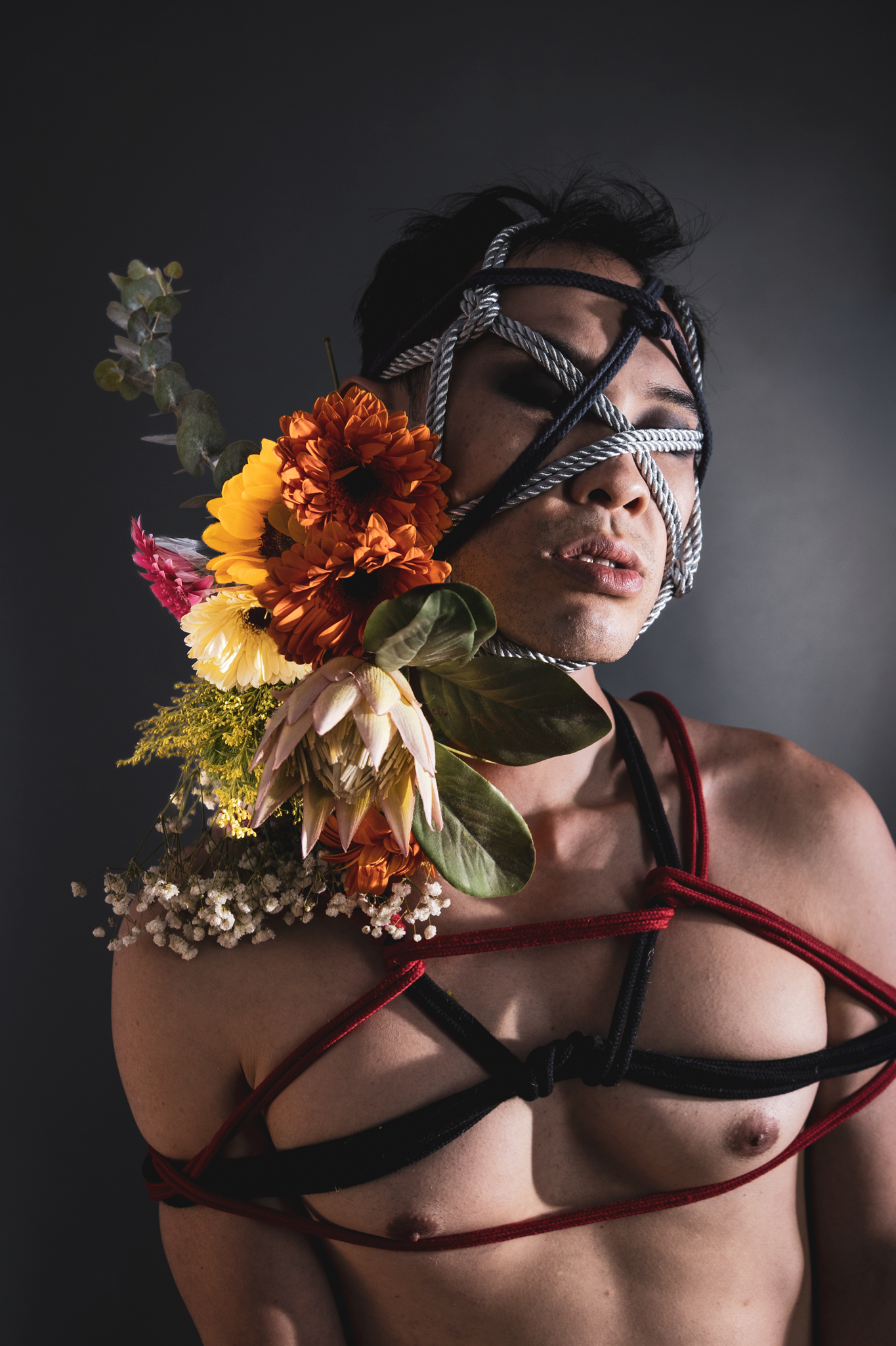 Artistic portrait of a person with rope across face and colorful flowers, wearing body harness in studio.