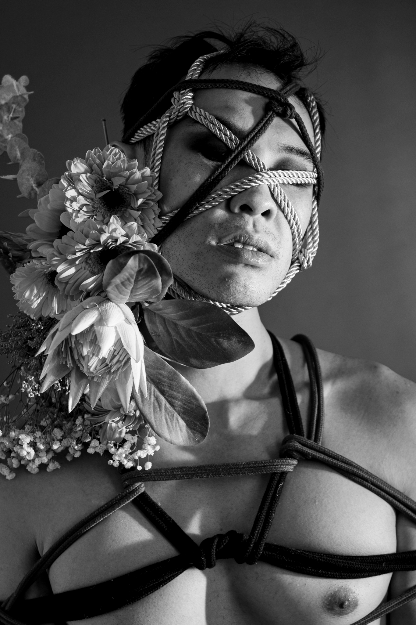 Black and white artistic portrait of a person with rope bondage on face and torso, decorated with flowers.